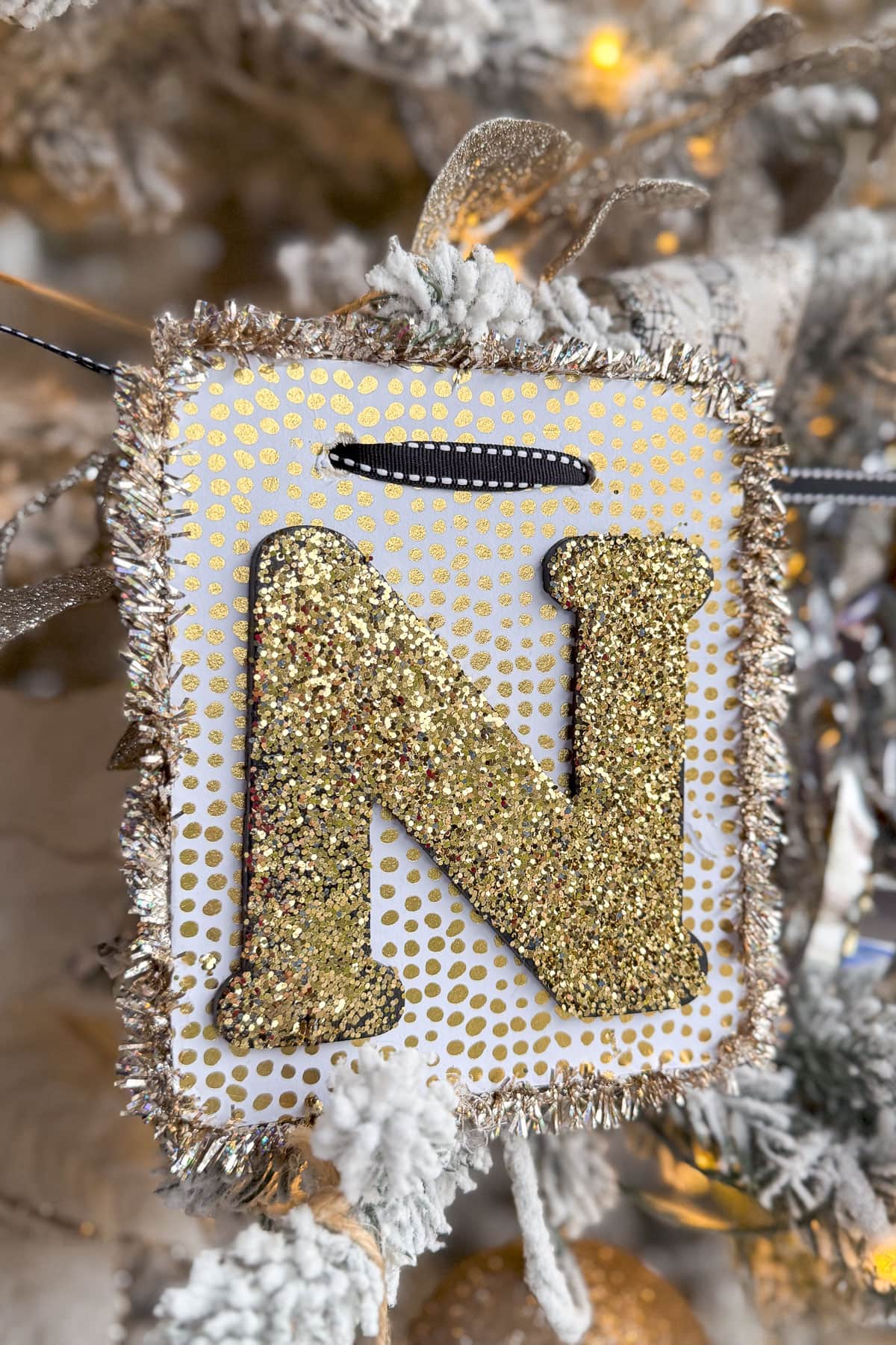 A festive ornament hanging on a Christmas tree features a glittery gold letter "N" against a white backdrop adorned with gold polka dots. The edges are trimmed with silver and gold tinsel, complemented by elegant New Year's Tree Decorations in white and silver.