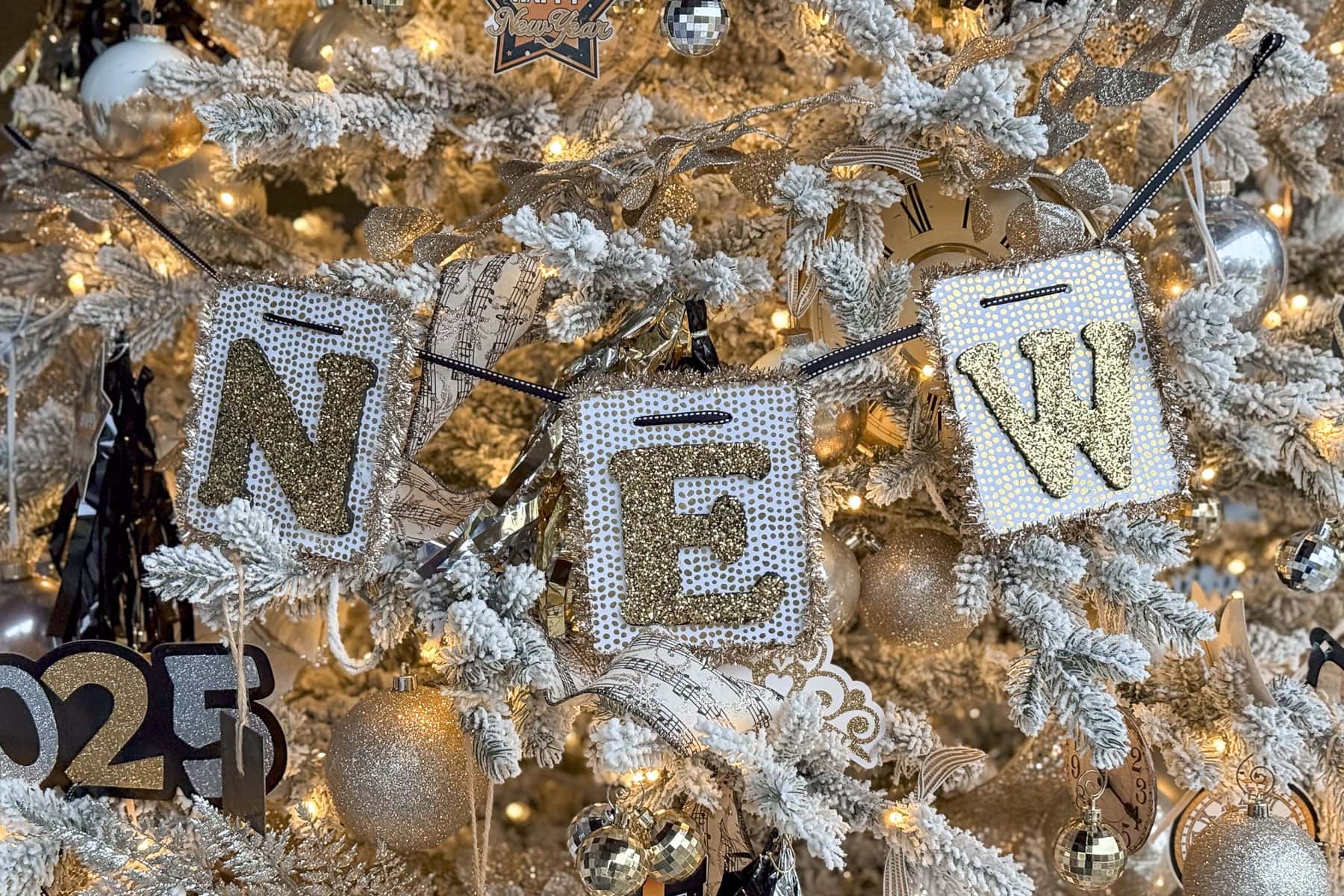 A festive New Years tree adorned with gold and white decorations, twinkling lights, and a sparkling  DIY banner spelling out "NEW" in sequined fabric. Ornaments include glittery balls and numbers "2025.