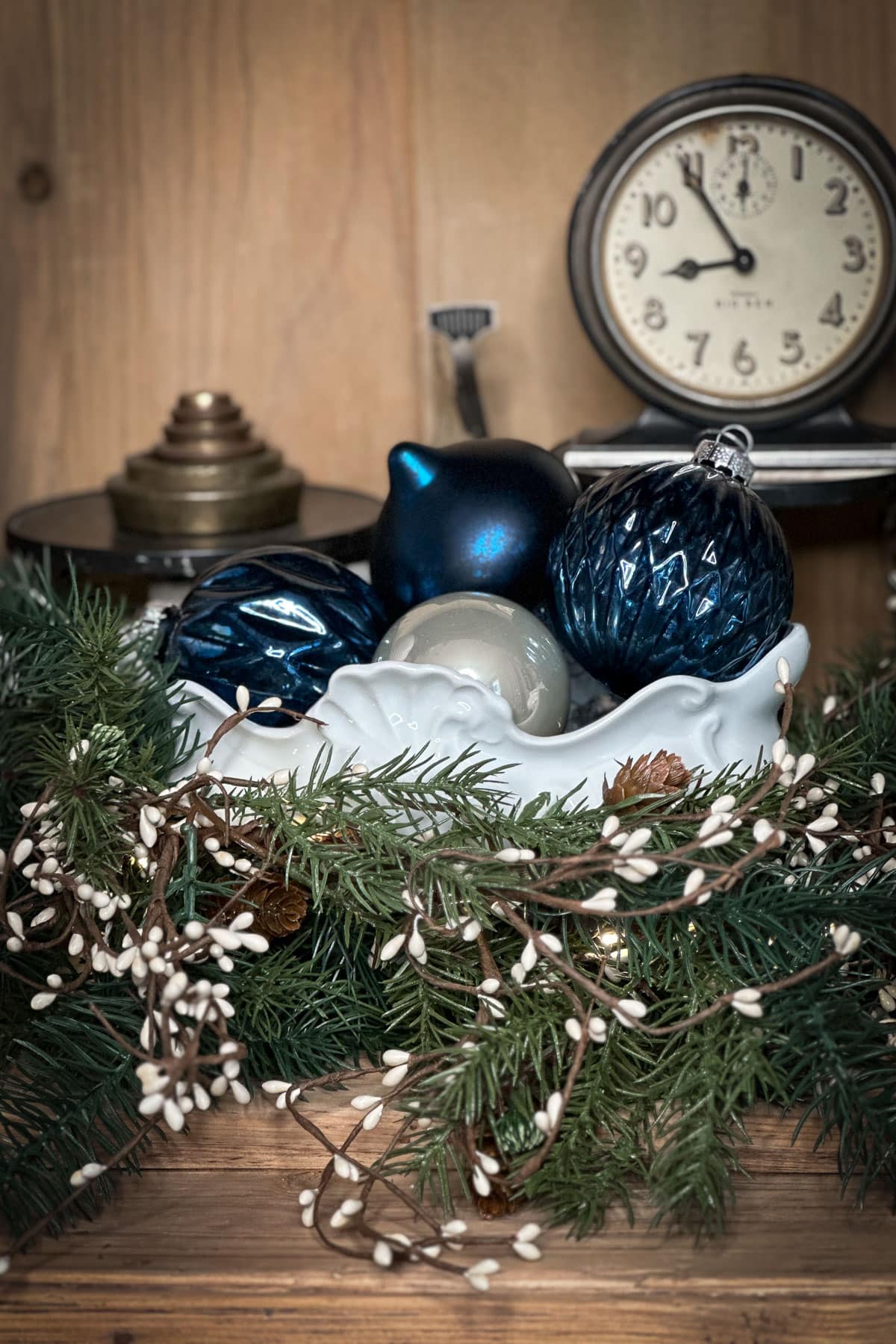 A classic Christmas decor scene showcases a white bowl filled with blue and silver ornaments, nestled among evergreen branches and white berry sprigs. A vintage clock and candle holder add charm to the wooden backdrop, embodying timeless holiday elegance.