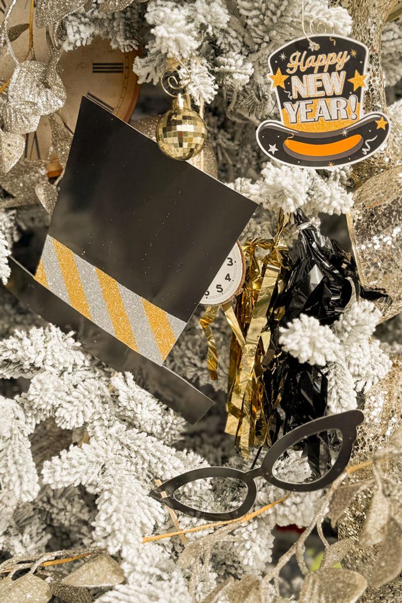 Close-up of a festive New Year's Eve decoration, featuring a frosted Christmas tree adorned with a "Happy New Year" sign, golden accents, a small clock, and black glasses. The tree is covered in white, snowy textures.