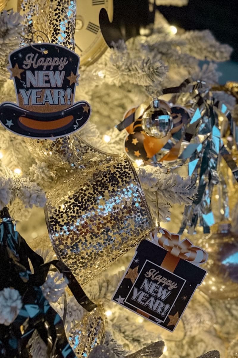 A close-up of a decorated New Year’s tree with gold and black ornaments. The tree features "Happy New Year" signs, shiny ribbons, a clock motif, and lit lights, creating a festive atmosphere.