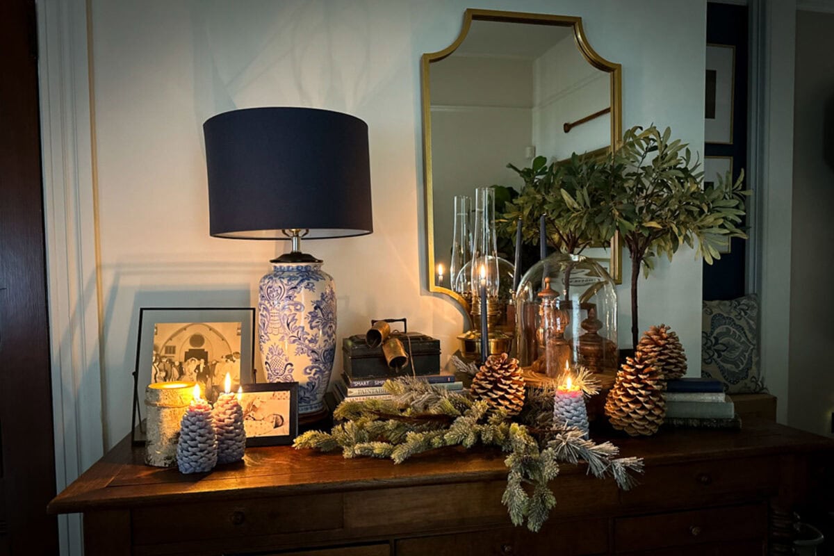 A cozy interior scene with a wooden dresser adorned with blue patterned lamp, pine cones, greenery, candles, and framed photos. A large mirror reflects the warm glow from the candles, creating an inviting and festive atmosphere.