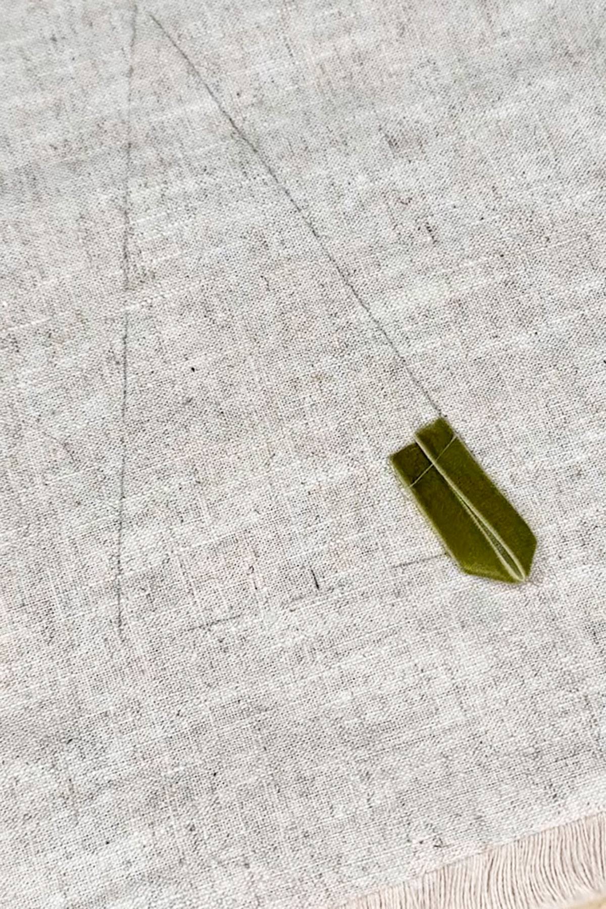 A piece of fabric with a faint triangle drawn on it. A small strip of green painter's tape is placed near the bottom of the triangle. The fabric has a textured surface and frayed edge at the bottom.