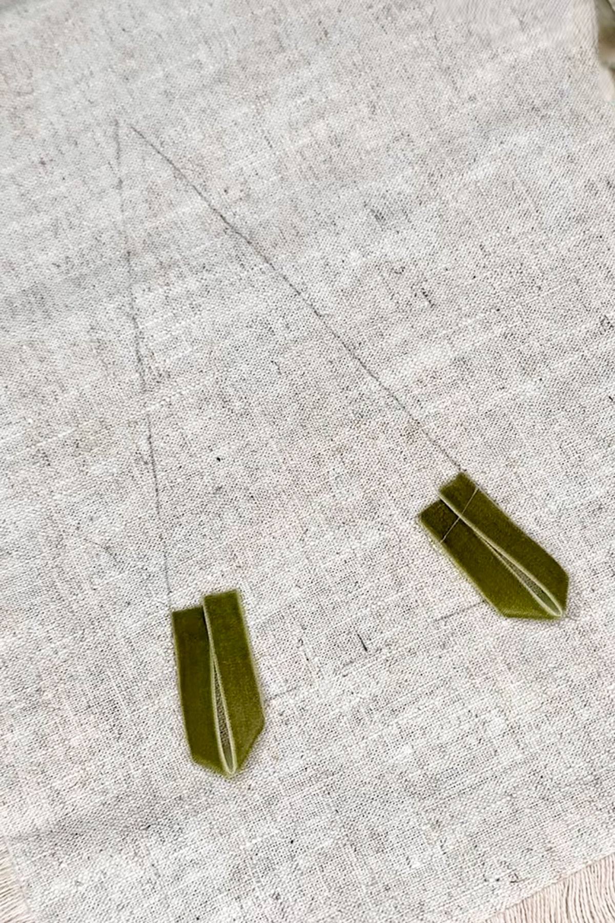 Two green origami cranes attached to strings lie on a textured beige surface. The strings are thin and barely visible against the fabric backdrop, creating a delicate and minimalist composition.