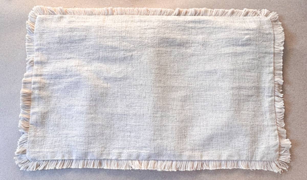 A rectangular beige cloth with fringed edges lies flat on a surface. The texture is coarse, resembling burlap, and it contrasts against the light background.