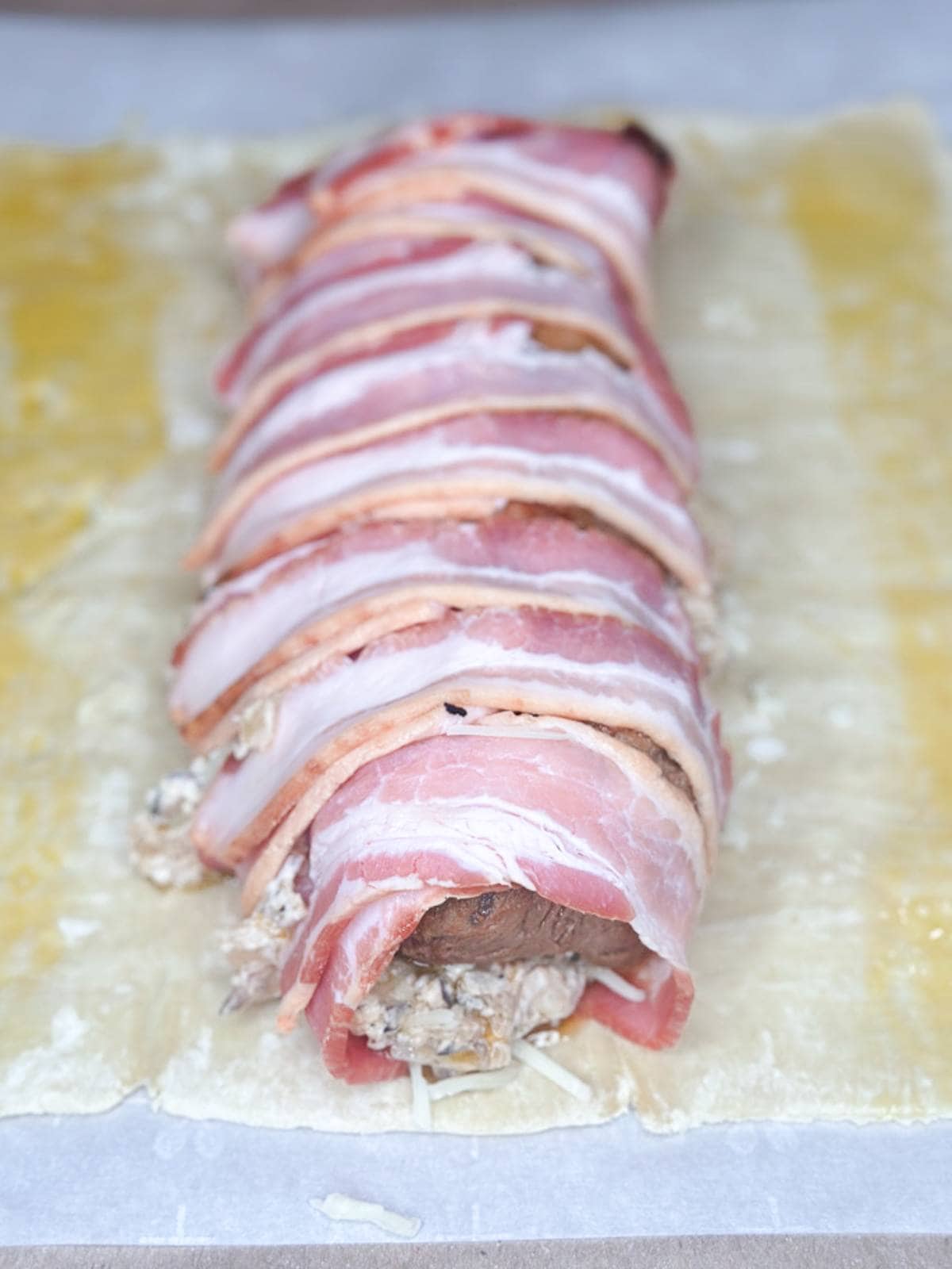 A raw beef Wellington preparation is shown. The beef is wrapped in prosciutto with a mushroom duxelles filling, all placed on a sheet of puff pastry. The pastry is unrolled, ready for wrapping and baking.