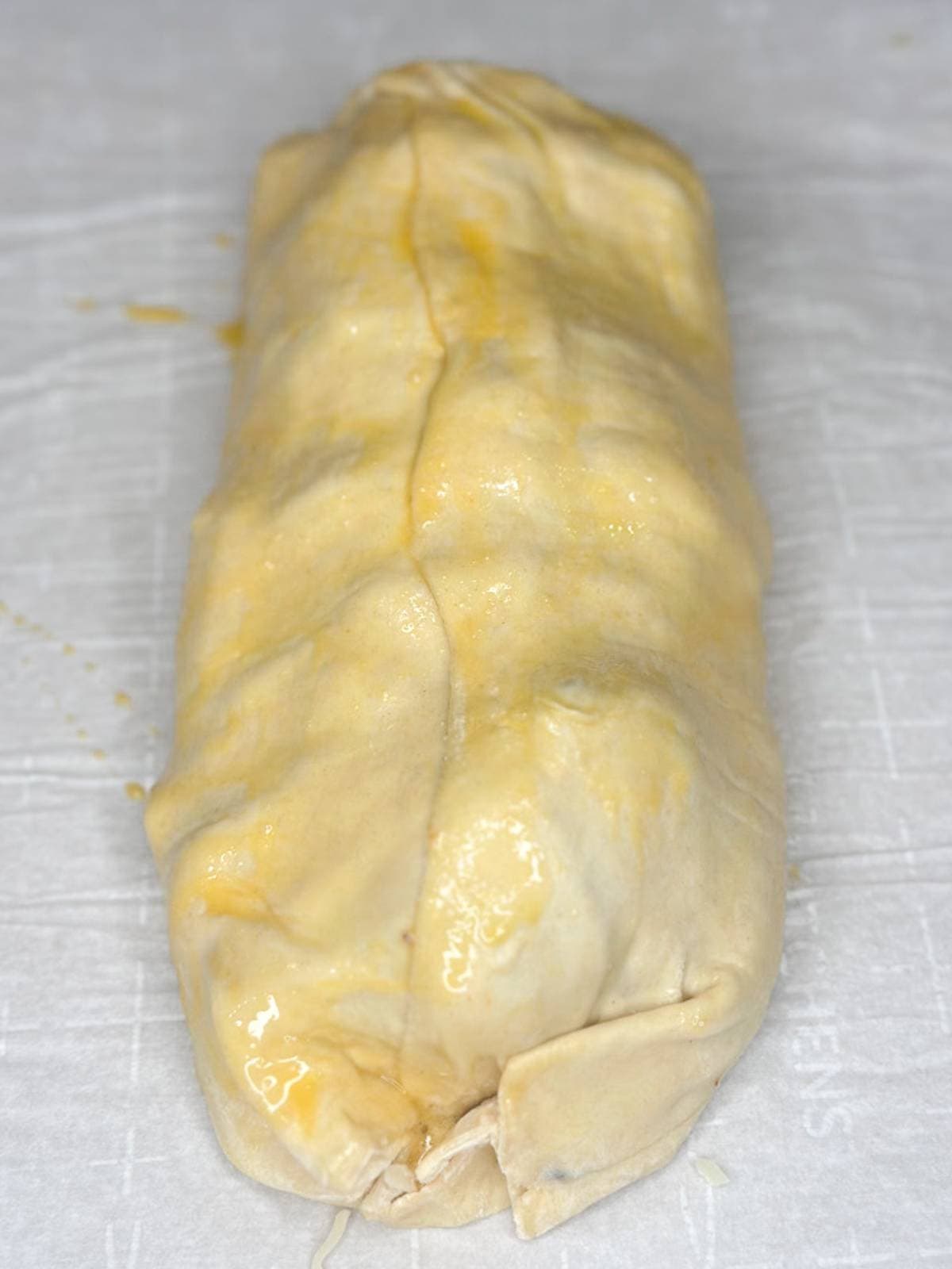 A close-up of an uncooked pastry roll, brushed with egg wash, on a white parchment paper. The pastry has a folded seam running down the middle and visible creases at each end.