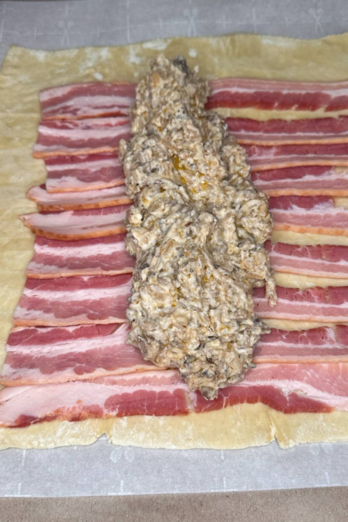 A rectangular sheet of dough is topped with evenly spaced strips of bacon. In the center, there's a mixture of creamy filling, possibly containing cheese and finely chopped ingredients, spread in a line down the middle.