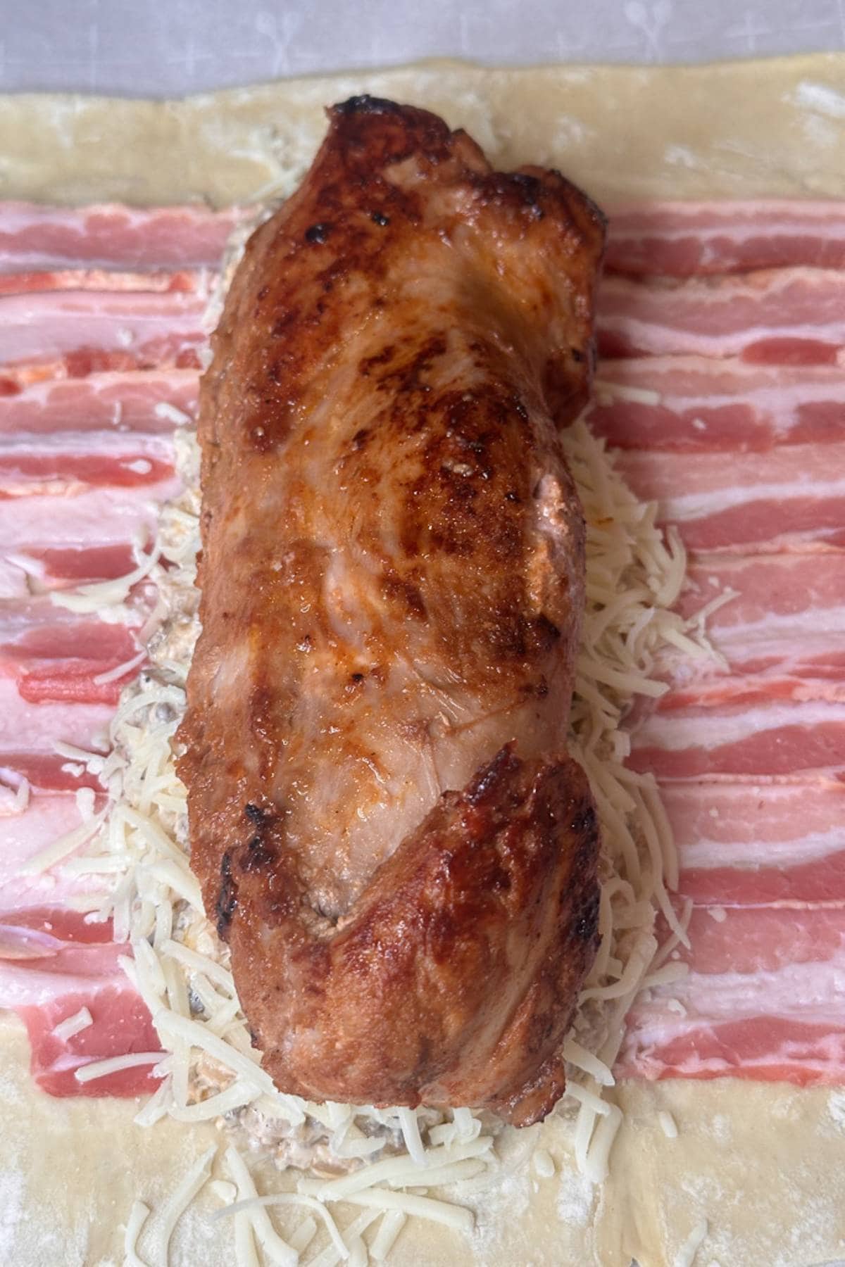 A browned piece of meat resting on a layer of grated cheese and strips of bacon, all placed on a sheet of dough.