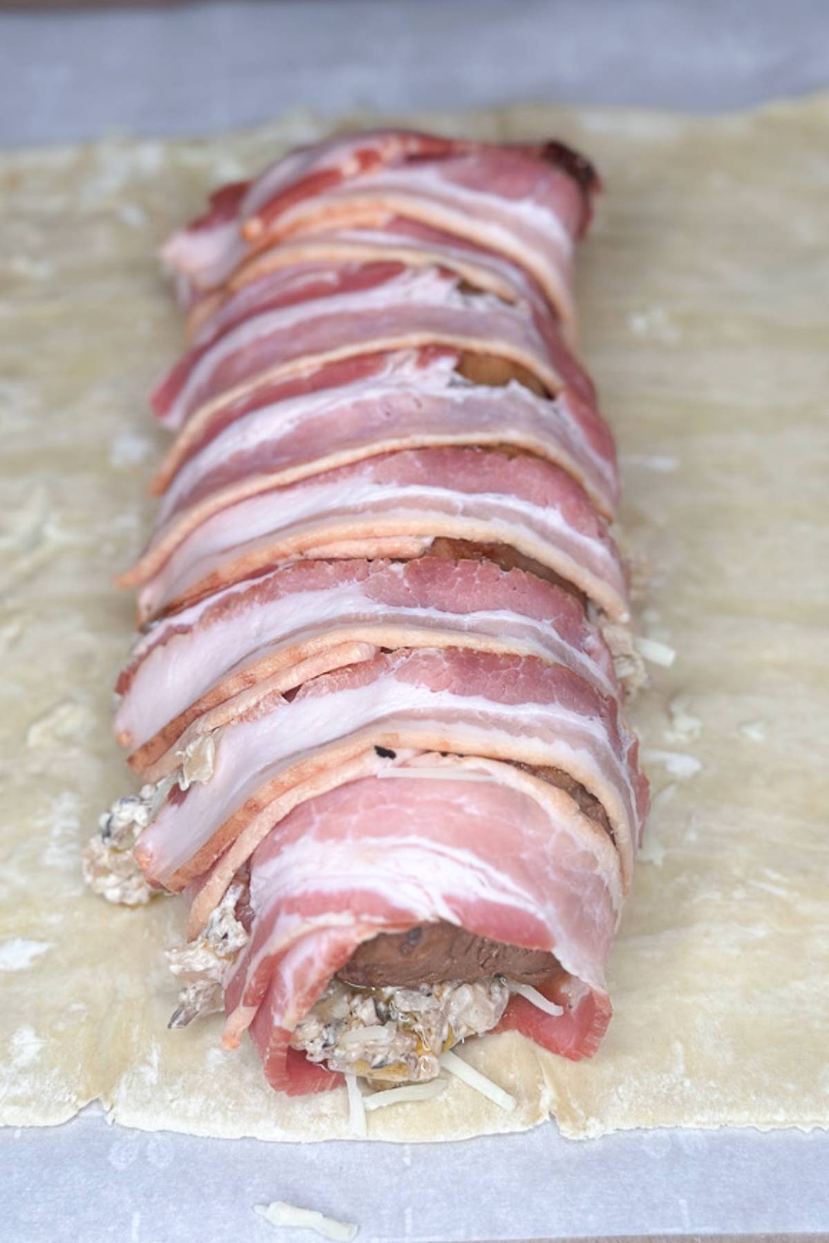 A rolled pastry on a baking sheet, wrapped in uncooked bacon strips. The filling includes visible mushrooms and cheese. The pastry dough is partially folded around the bacon and filling, ready to be baked.