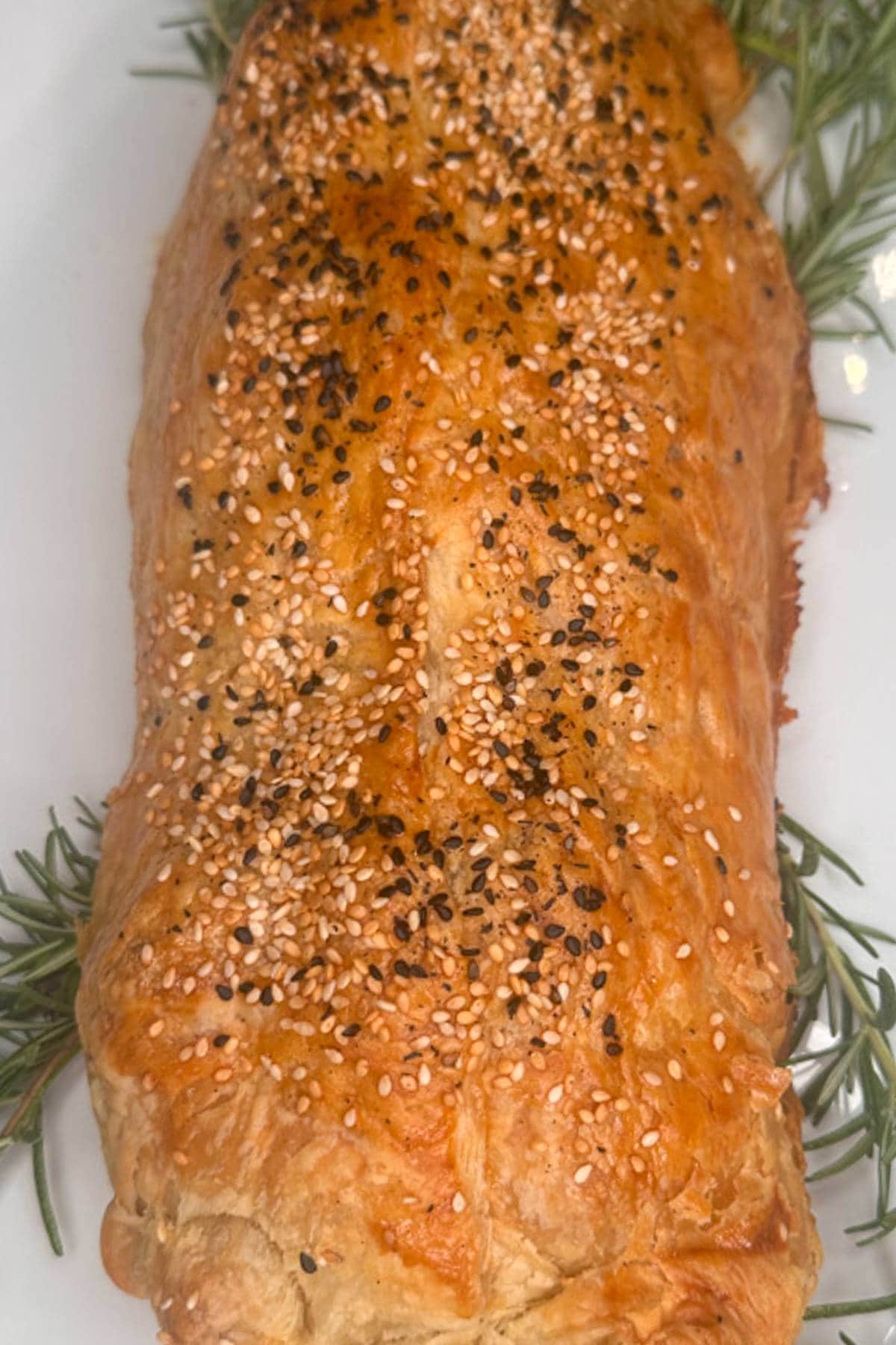 Golden-brown puff pastry loaf sprinkled with sesame and black seeds, garnished with sprigs of fresh rosemary on a white plate.