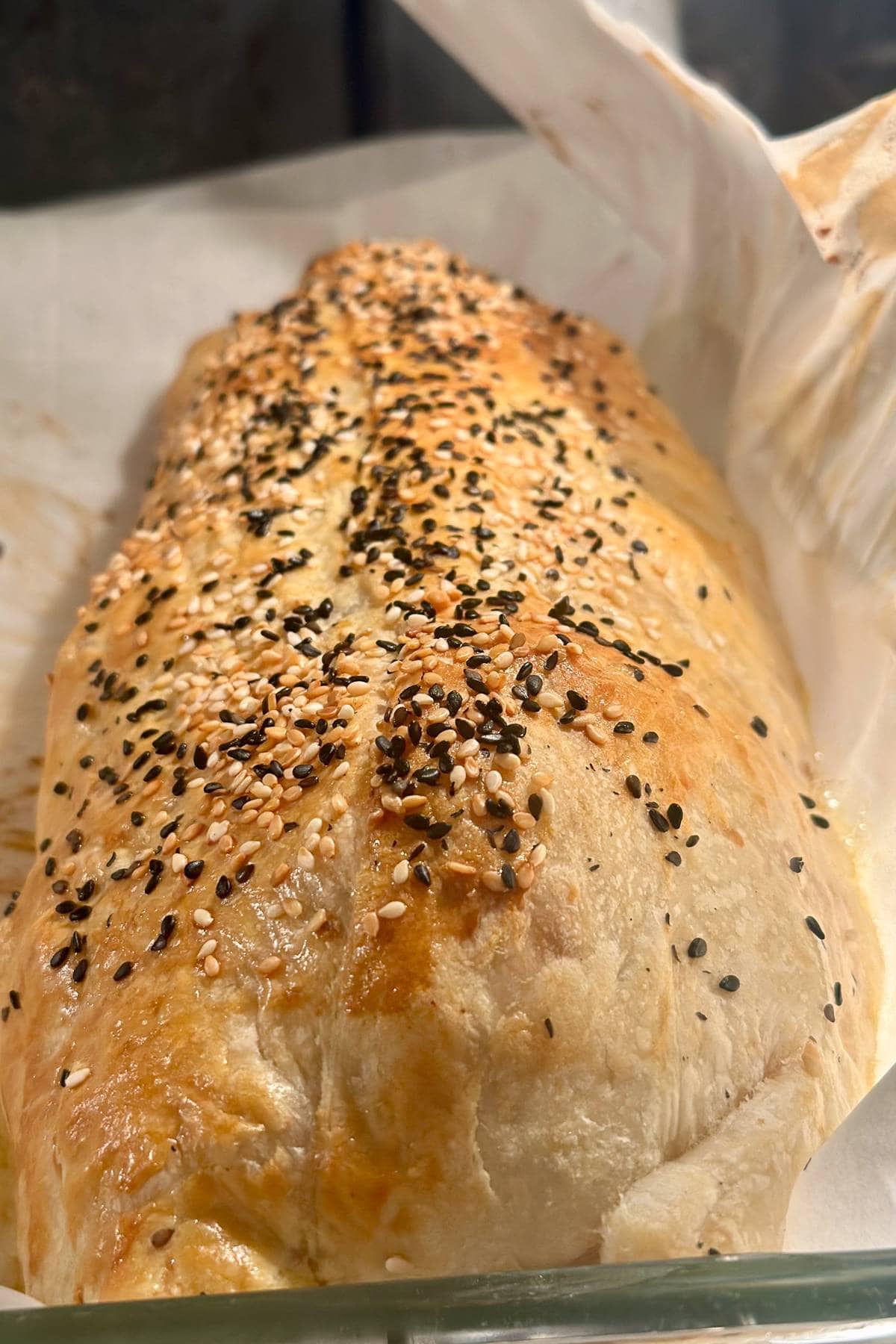 A close-up of a freshly baked loaf of bread topped with a generous sprinkle of black and white sesame seeds, resting on parchment paper. The bread has a golden-brown crust and looks warm and inviting.