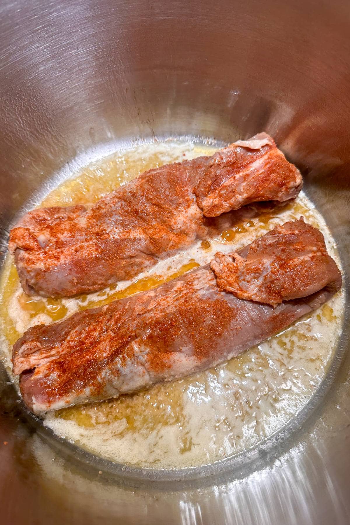 Two seasoned pork tenderloins searing in a hot pan with a layer of melted butter, showing a golden brown crust. The seasoning is visibly red, indicating spices like paprika or cayenne. The pan is metallic with slight oil splatters around the meat.