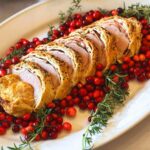 Sliced Pork Wellington garnished with fresh rosemary and cranberries on a white platter. The meat is encased in golden pastry, surrounded by vibrant red berries and green herbs.