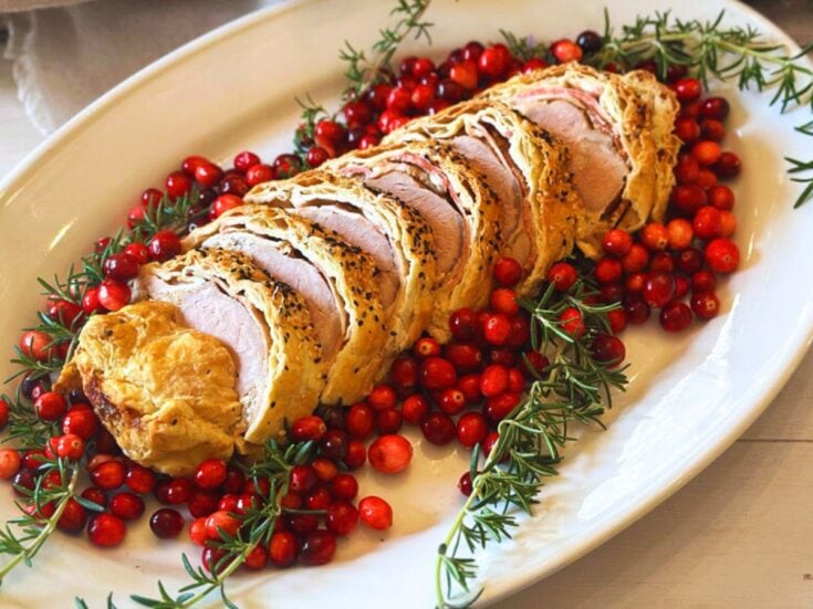 Sliced Pork Wellington garnished with fresh rosemary and cranberries on a white platter. The meat is encased in golden pastry, surrounded by vibrant red berries and green herbs.