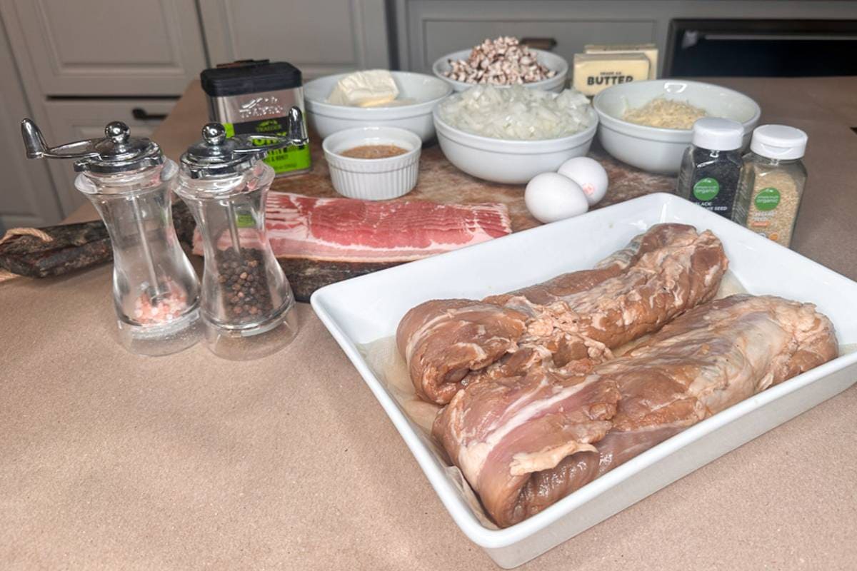 A kitchen counter with ingredients for a  pork wellington recipe, including two raw pork tenderloins in a white dish, pepper, salt, eggs, bacon, chopped onions, mushrooms, butter, and spices. Everything is ready for cooking.