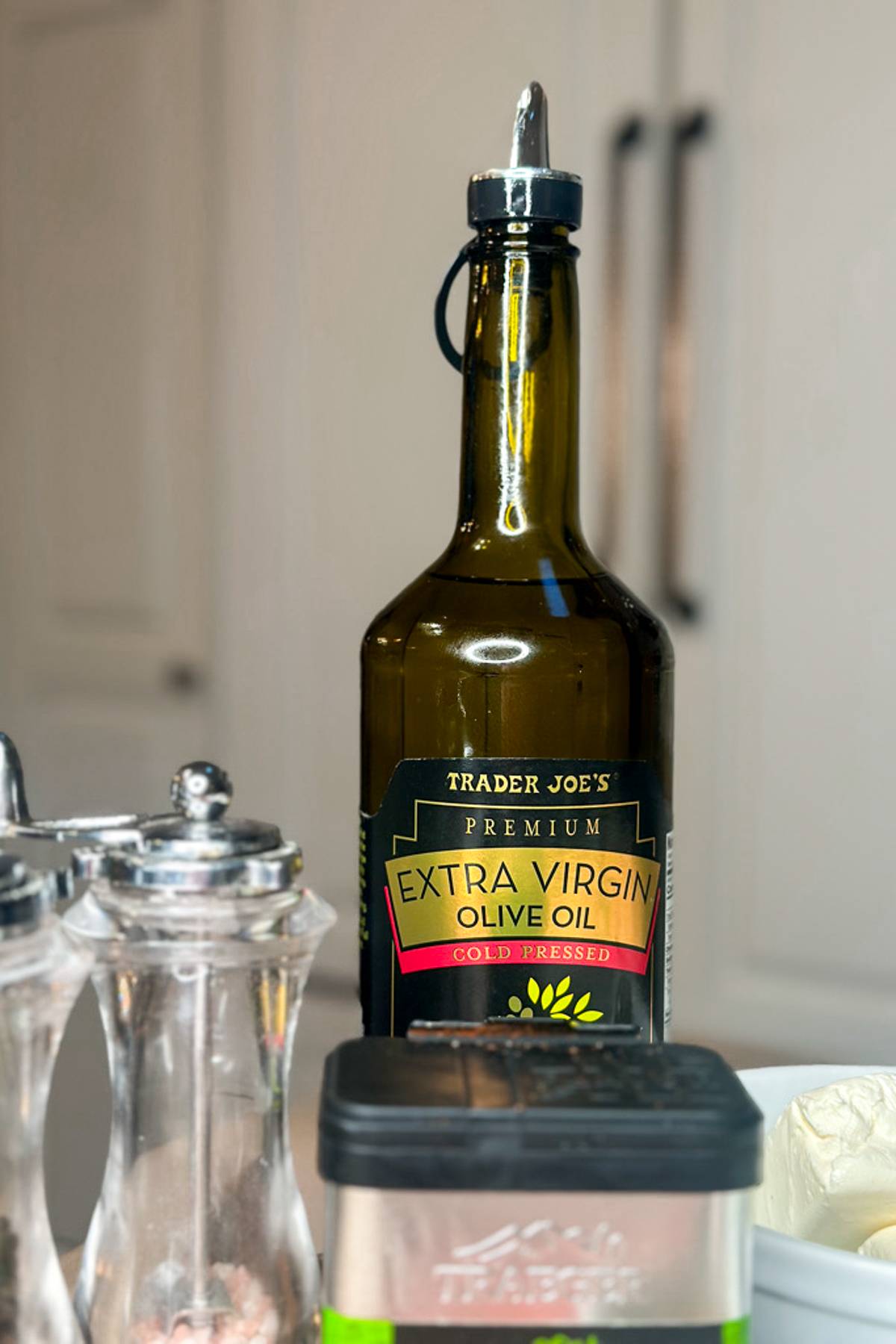 A bottle of Trader Joe's premium extra virgin olive oil is placed on a kitchen counter. Next to it are salt and pepper shakers and a container with a silver lid. The background is a blurred white kitchen cabinet.