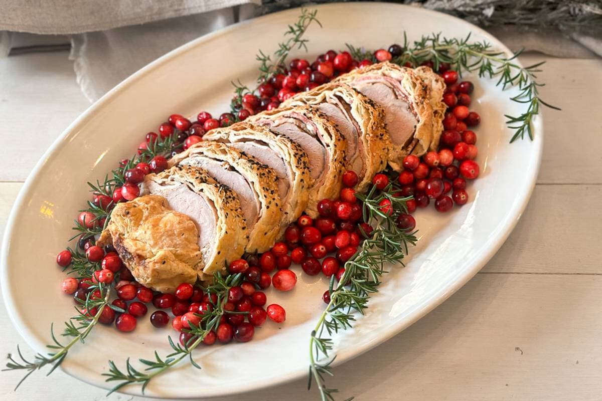 A sliced meat dish wrapped in pastry is displayed on an oval platter. It is garnished with fresh cranberries and sprigs of rosemary, creating a festive presentation.