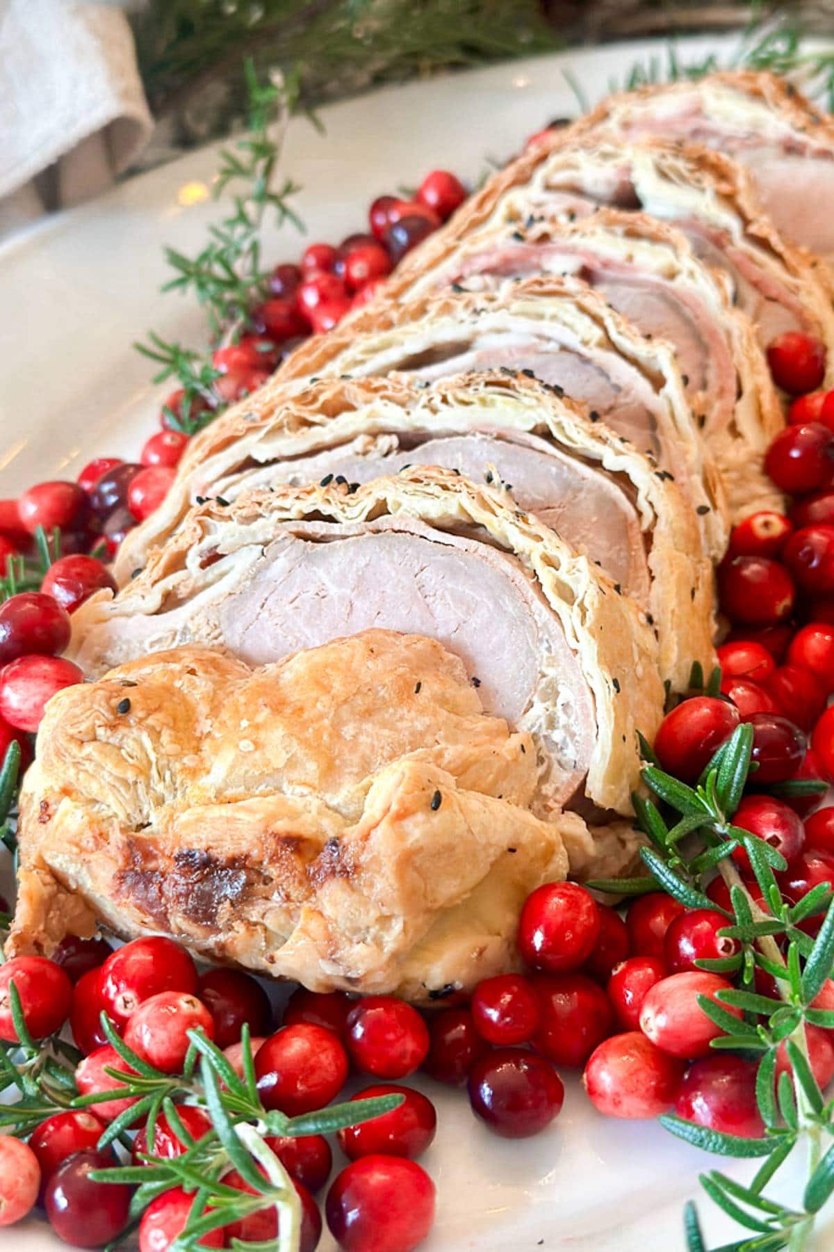 Sliced pork loin wrapped in flaky pastry, garnished with fresh cranberries and rosemary sprigs on a white plate.