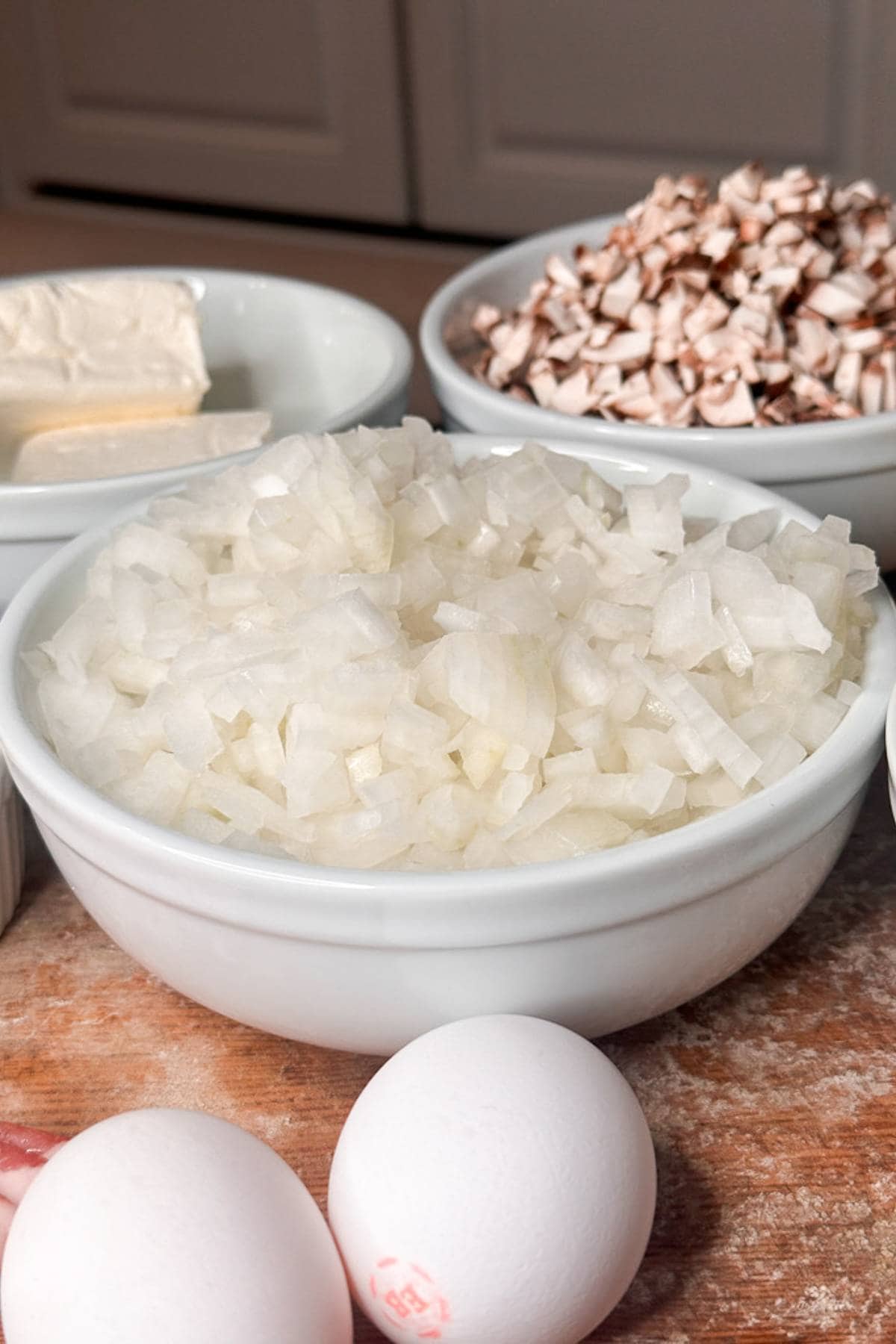 A close-up of a kitchen counter with a bowl of chopped onions in focus. Behind are bowls of diced mushrooms and butter. Two whole eggs are in the foreground.