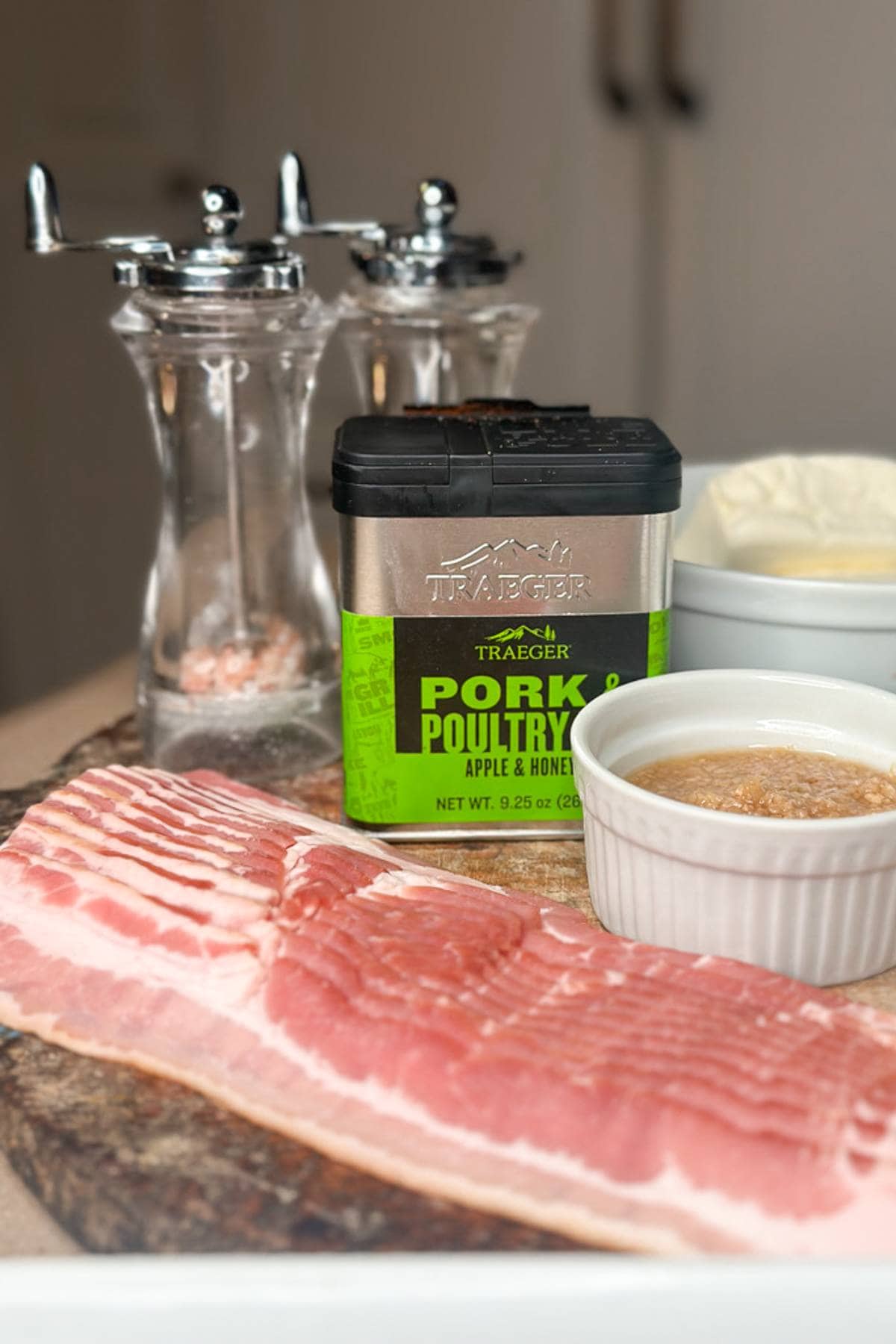 Sliced bacon on a cutting board next to a small bowl of brown liquid, a green container of Traeger pork and poultry rub, and two salt and pepper grinders.