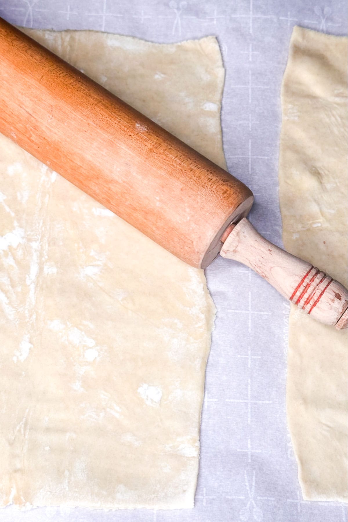 Rolling pin on a sheet of uncooked, light-colored pastry dough awaits its transformation into a savory Pork Wellington. Laid out on a lightly floured surface, the dough is partially rolled with some uneven edges, ready to envelop the flavorful filling.