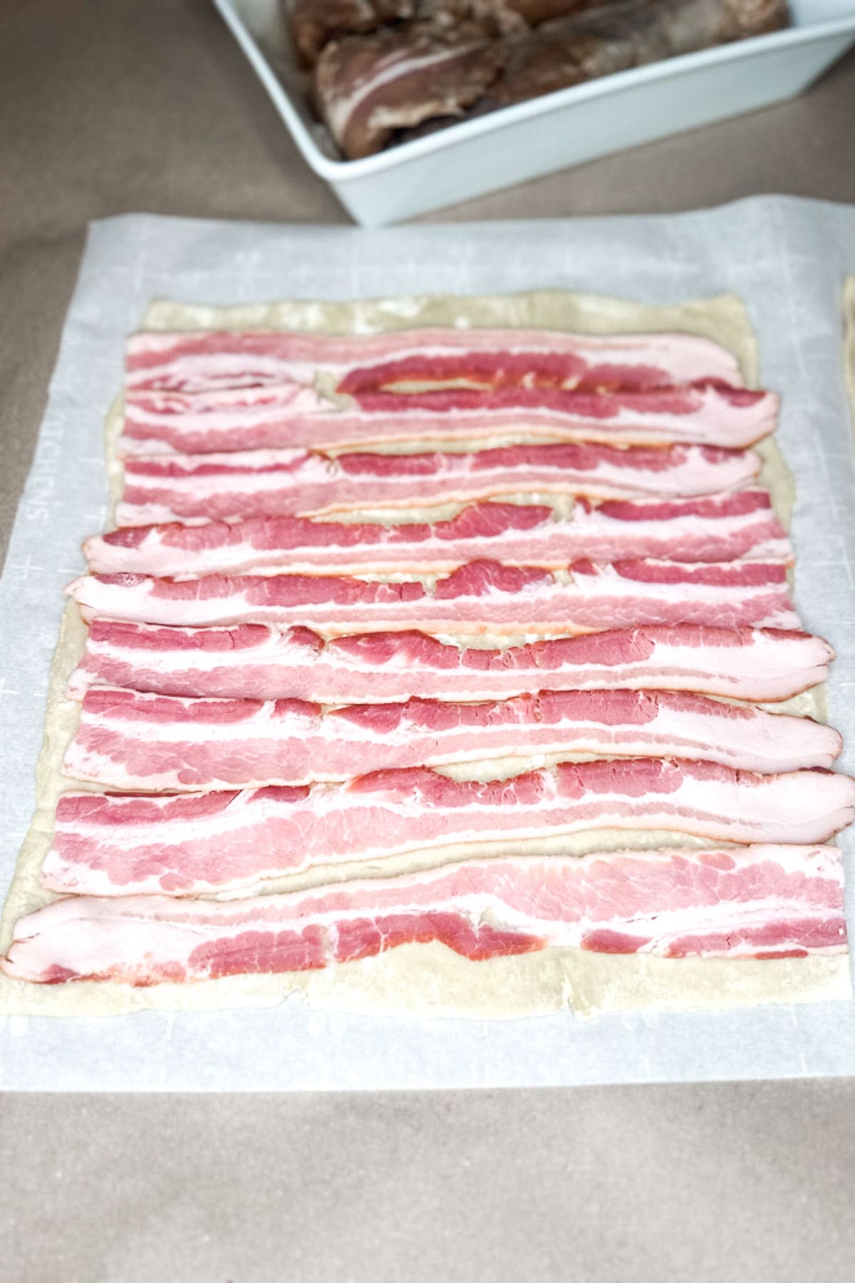 Slices of raw bacon are arranged in a neat grid pattern on a piece of puff pastry on top of a white parchment paper, ideal for wrapping a savory Pork Wellington. In the background, a white tray holds more raw meat, ready to be transformed into culinary art.