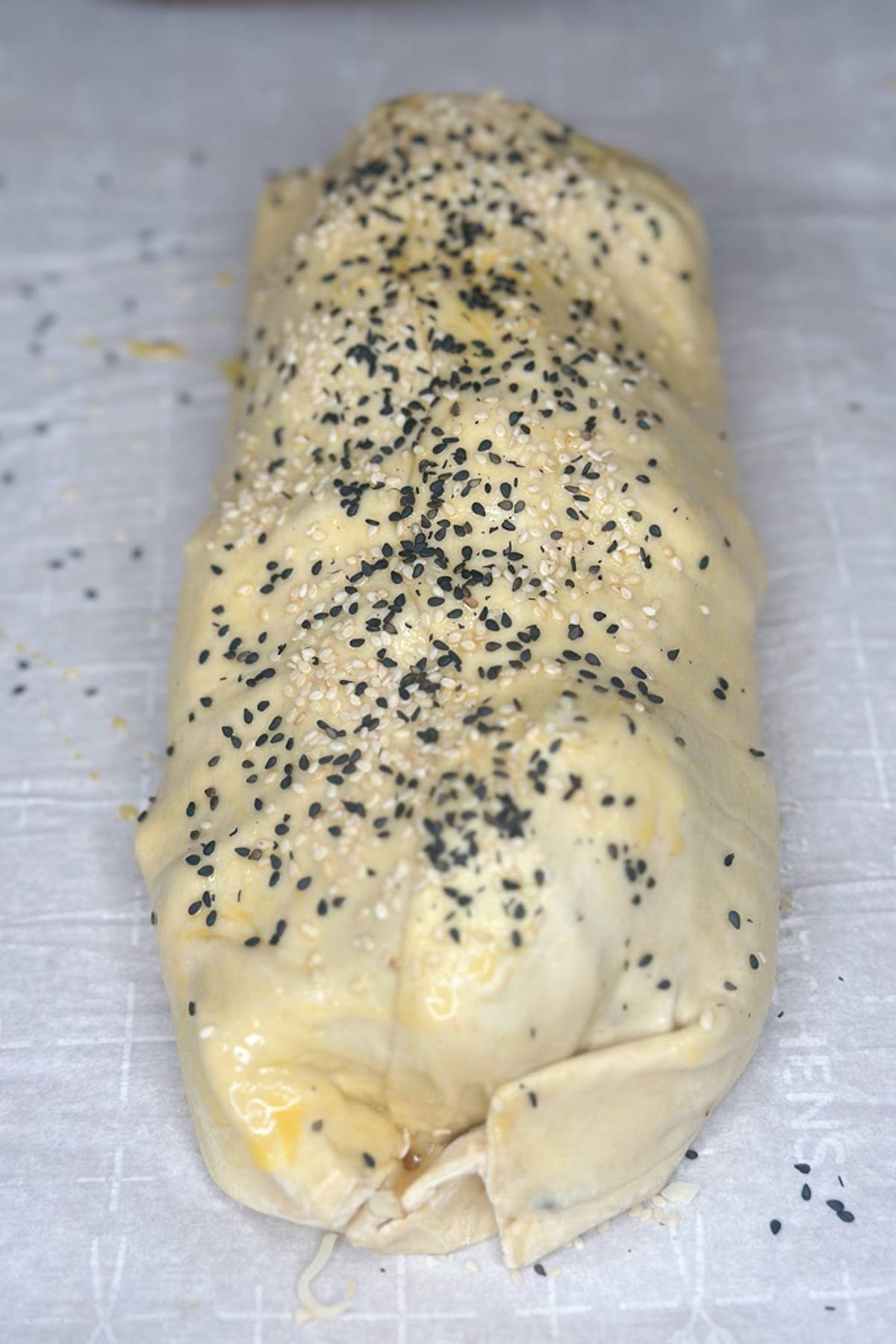 A partially baked pastry roll on parchment paper, sprinkled with black and white sesame seeds. The dough is golden and slightly puffed, hinting at a filling inside.