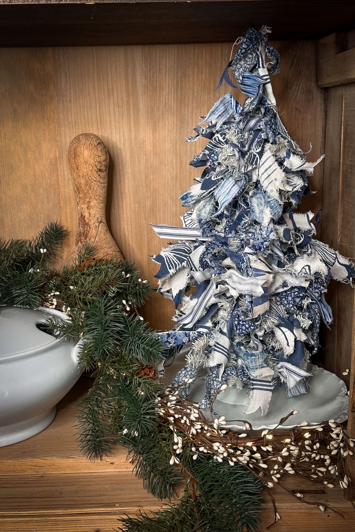 A small fabric Christmas tree made of blue and white strips sits on a wooden shelf, embodying classic Christmas decor. Surrounded by green pine branches and a white ceramic bowl, a wooden rolling pin leans against the back, adding charm to the festive scene.