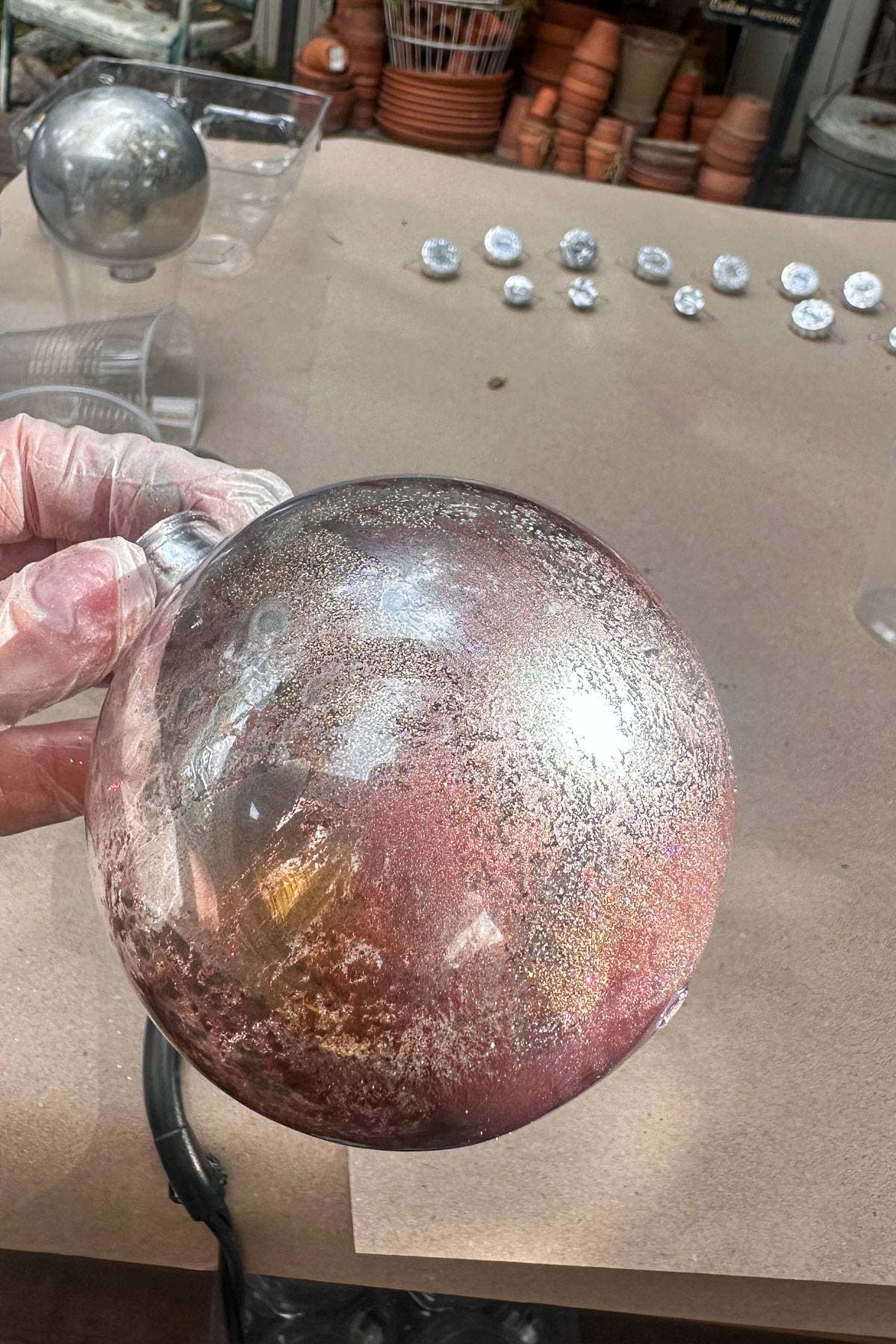 A gloved hand holds a large, round glass ornament with a metallic and speckled finish. In the background, there are several smaller ornaments and stacked terracotta pots on a table.