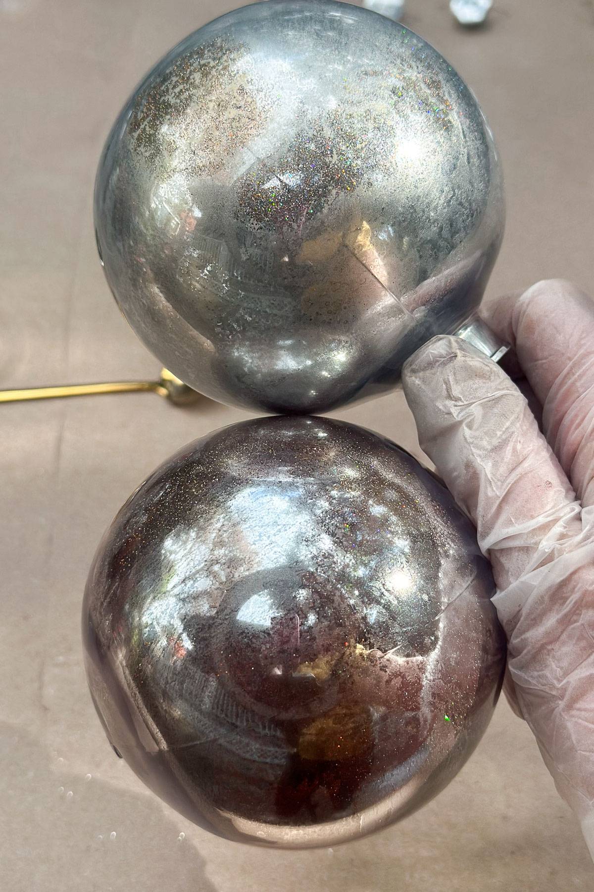 A gloved hand holding two metallic, reflective spheres. The spheres have a shiny surface with a mix of silver and bronze hues, capturing reflections of the surroundings. They are placed on a neutral, light-colored background.