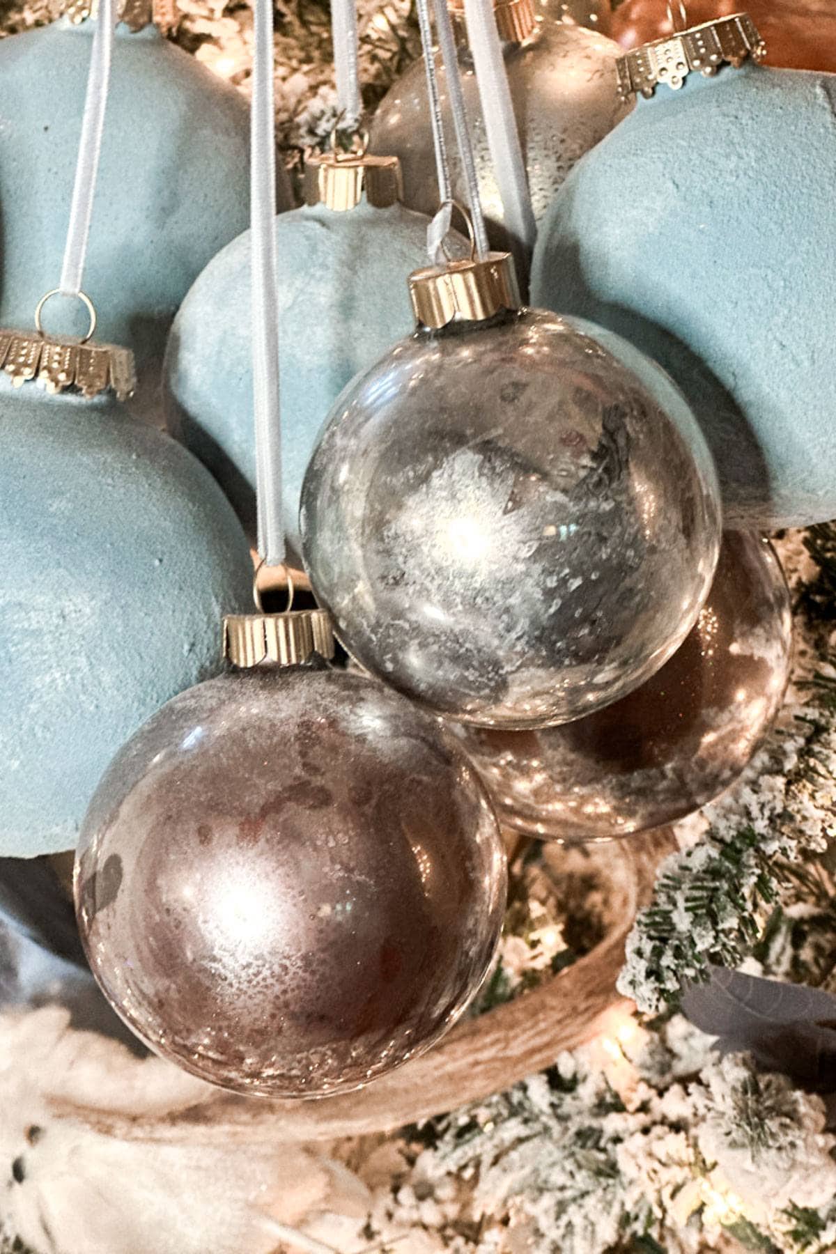 Close-up of Christmas tree ornaments, featuring shiny silver and matte blue baubles hanging from white ribbons. The tree is adorned with snow-like flocking and soft white lights, creating a festive and cozy holiday atmosphere.