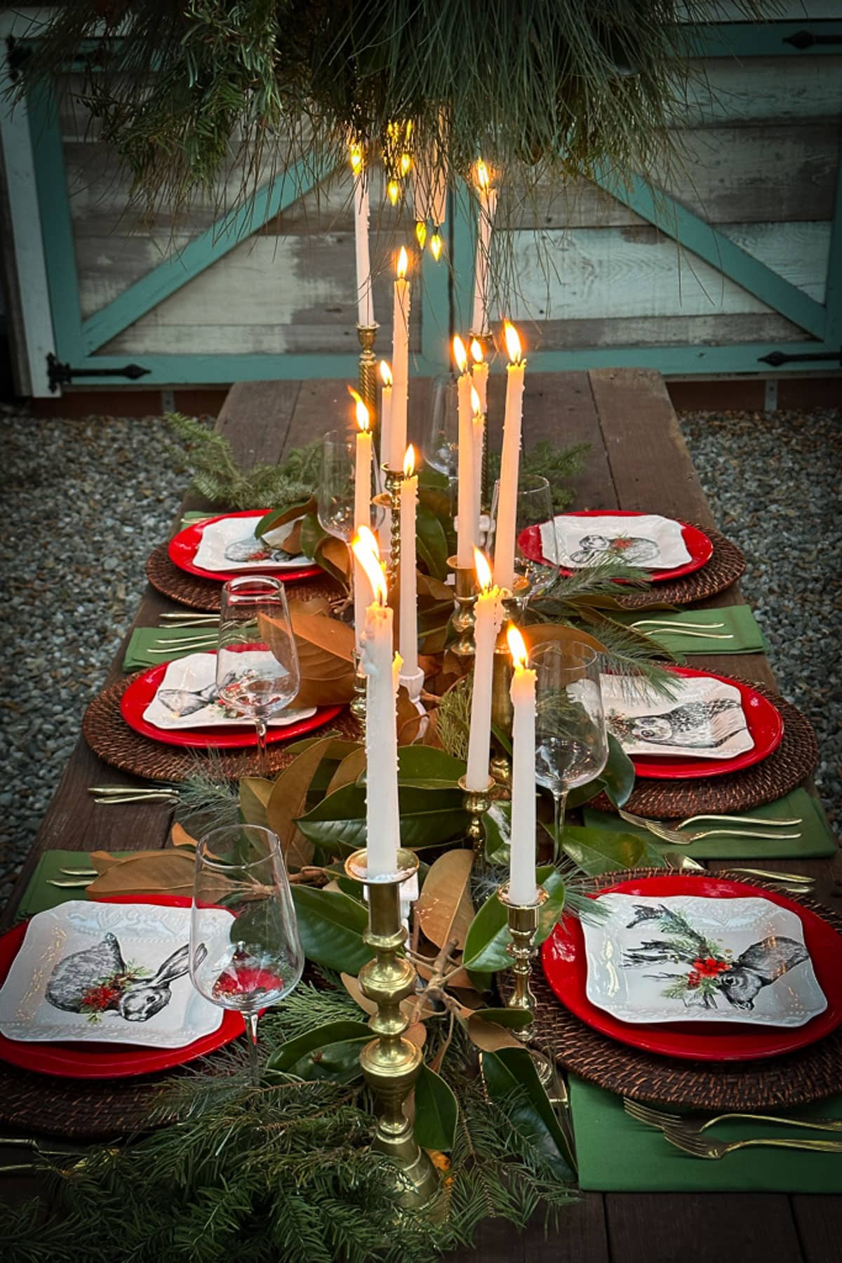 A rustic outdoor dining table is set for six with red and white plates featuring bird designs, surrounded by greenery and tall, lit candles. Wicker chargers and wine glasses add elegance, while a wooden barn door provides a charming backdrop.