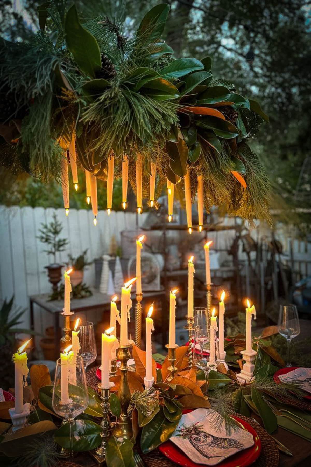 A beautifully set outdoor table features a lush greenery centerpiece with hanging taper candles and additional candles on the table. Plates, glasses, and napkins are elegantly arranged, creating a warm and inviting atmosphere.