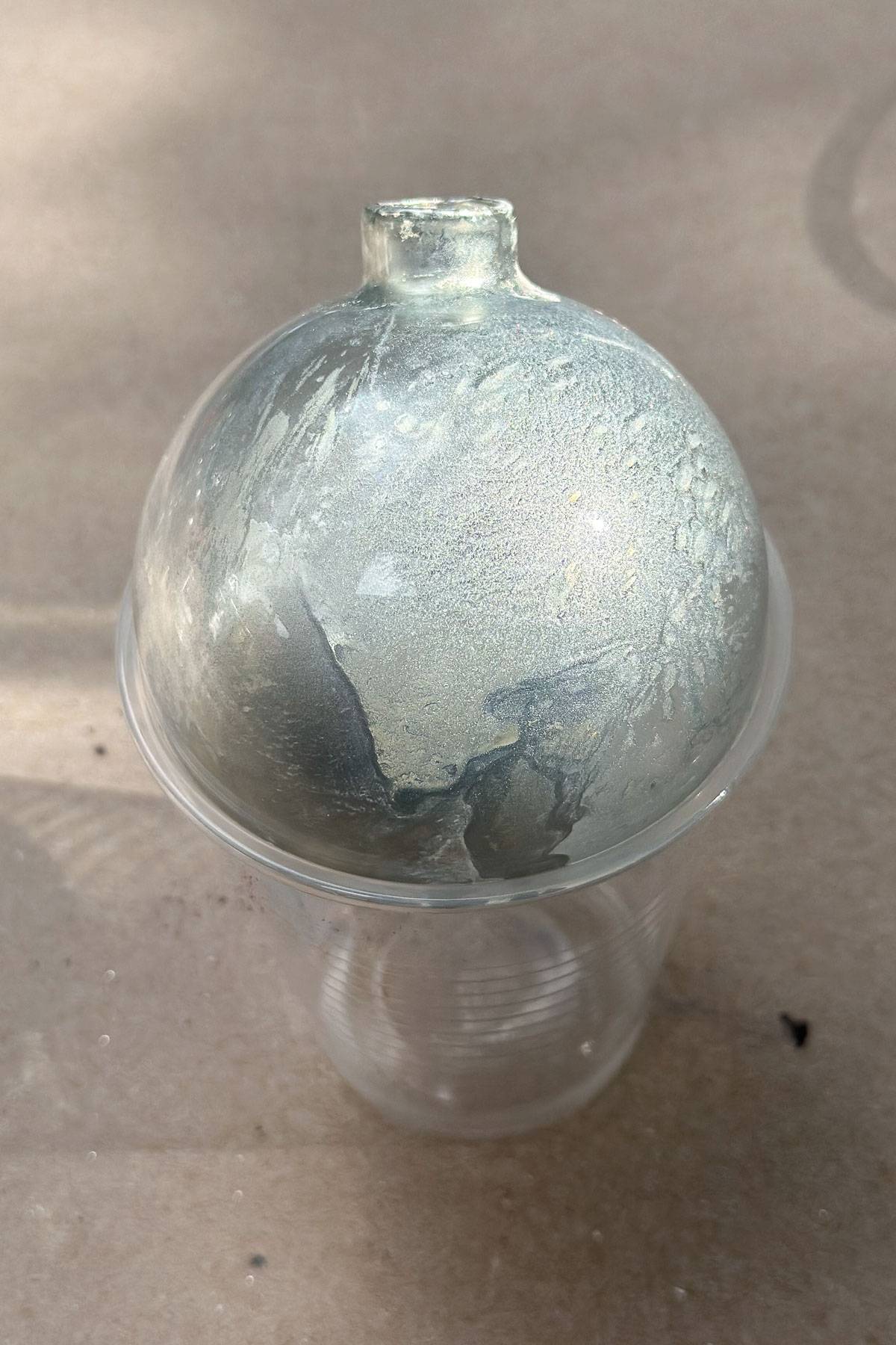 A translucent, frosted glass-looking ball positioned upside-down in a clear plastic cup. The surface has a textured, swirled pattern with shades of silver and gray, giving it a glossy appearance.