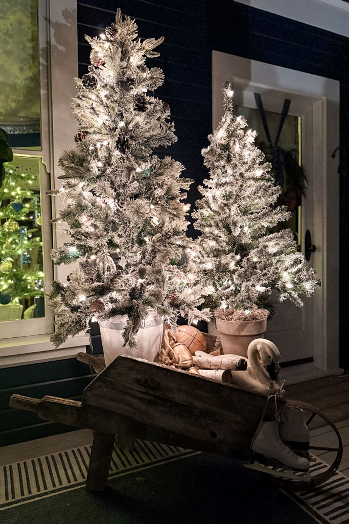 Two artificial Christmas trees with white lights stand in pots outside a house. A wooden wheelbarrow in the foreground holds white ice skates and a decorative goose, enhancing the festive, wintry atmosphere.