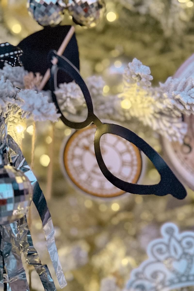 Festive decorations on a Christmas tree include black glasses, a disco ball ornament, and a clockface ornament. The tree is adorned with warm white lights and silver tinsel.