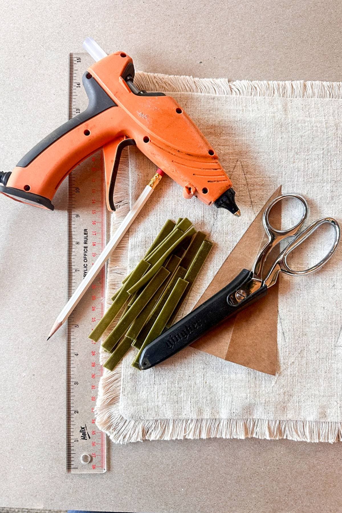 A crafting setup includes an orange hot glue gun, a pile of green glue sticks, a pair of scissors, a ruler, and a piece of triangular cardboard on a fringed fabric background.