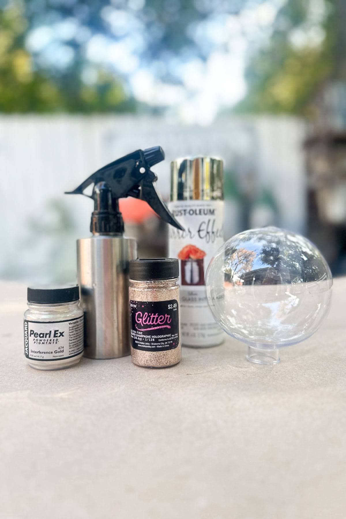 A collection of craft supplies on a table: a metallic spray bottle, glitter jar, Pearl Ex powder, silver effect spray can, and a clear plastic globe. Greenery and a white fence are blurred in the background.