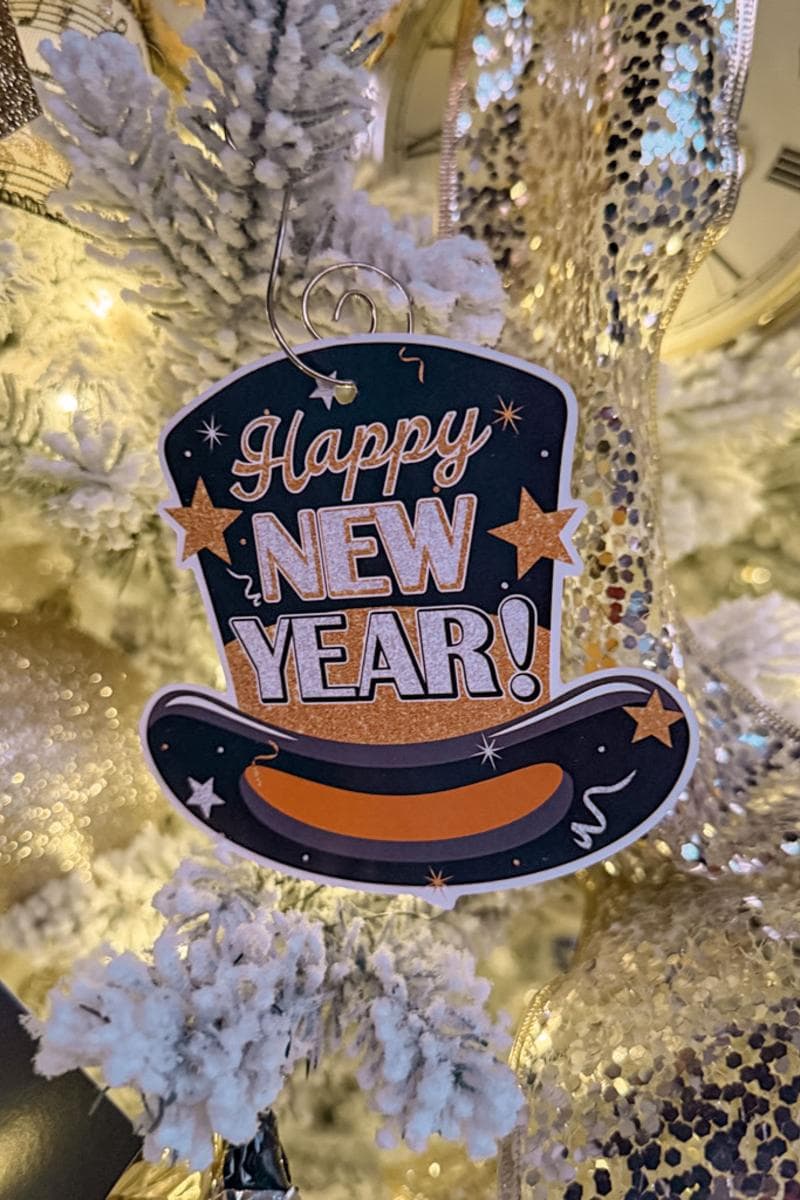 A festive decoration on a Christmas tree reads "Happy New Year!" in glittery gold and white letters on a black top hat shaped ornament, with stars and a shiny ribbon-adorned tree in the background.