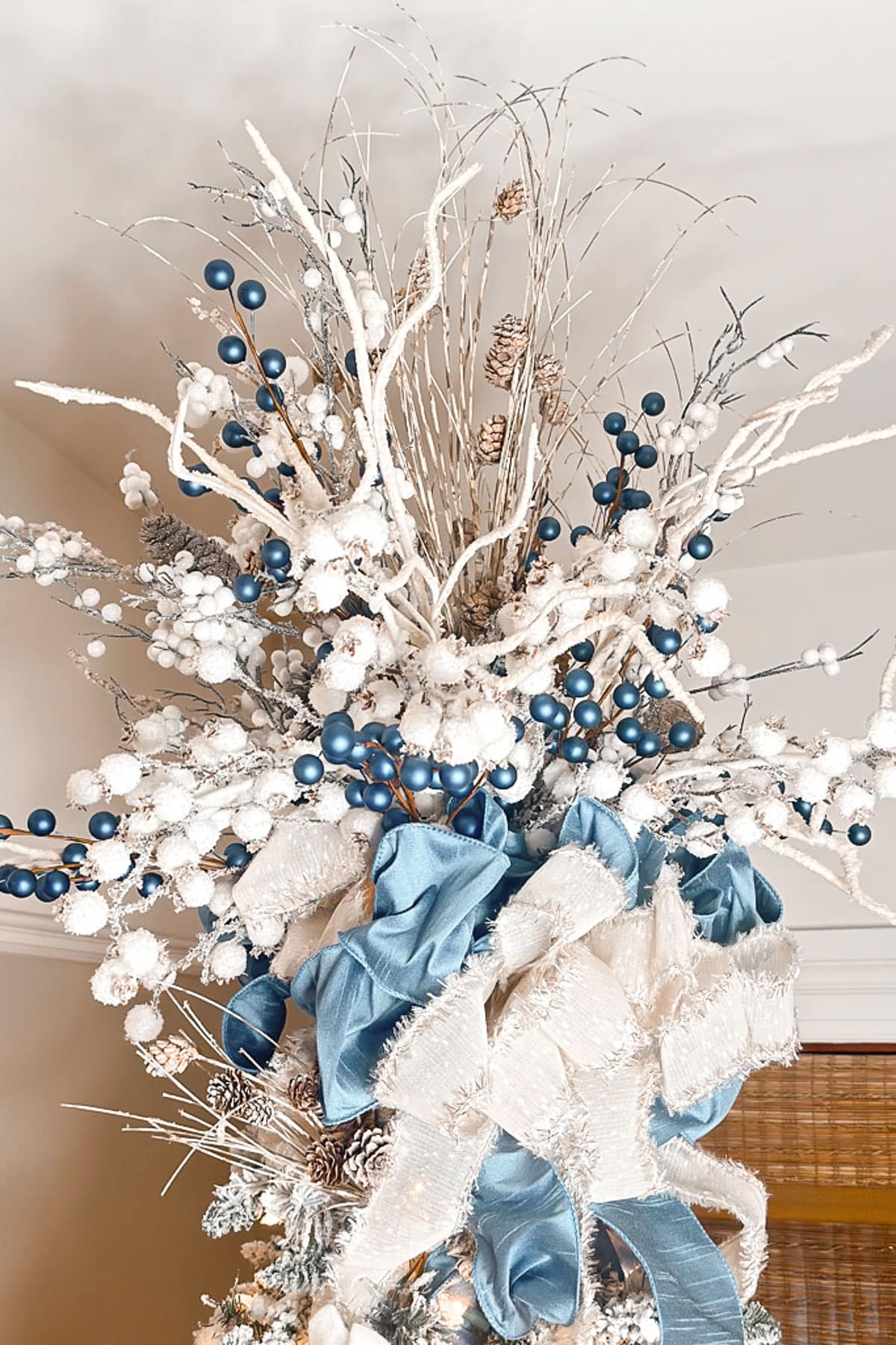 A festive, wintry floral arrangement featuring frosted branches with white and blue berries, white flowers, pine cones, and large blue and white ribbons, set against a neutral background.