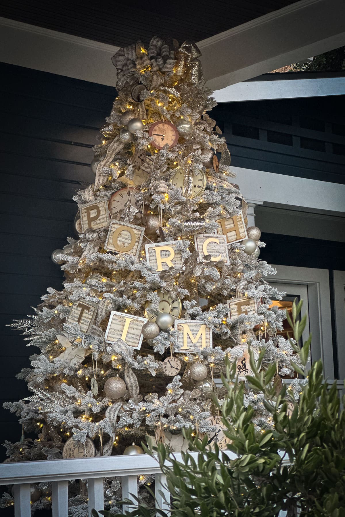 A tall, silver Christmas tree adorned with white and gold ornaments, clocks, and large block letters spelling "PORCH TIME." The tree is on a porch with dark walls and a white railing, and is illuminated with warm lights.