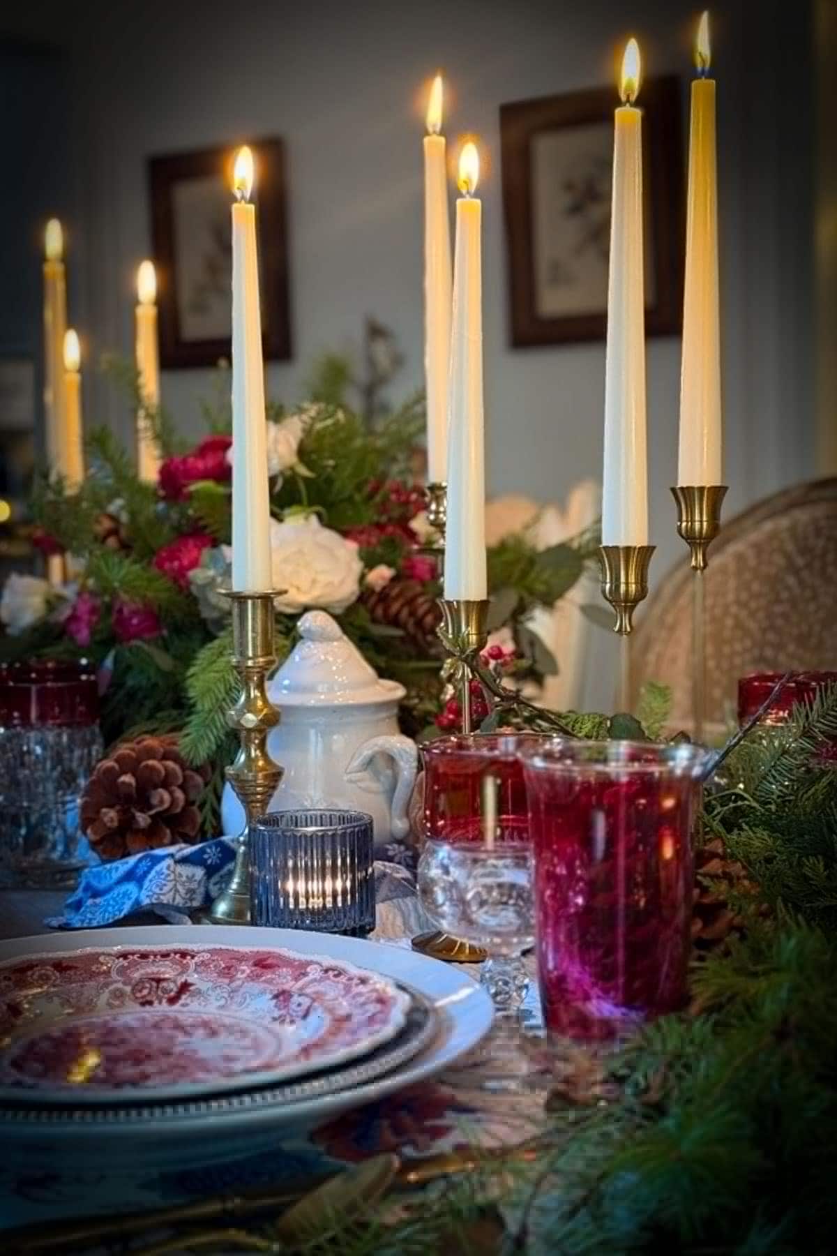 A festive dining table decorated with lit candles, red and white china, and pine greenery. The centerpiece features flowers and a pitcher, creating a cozy and elegant holiday atmosphere.