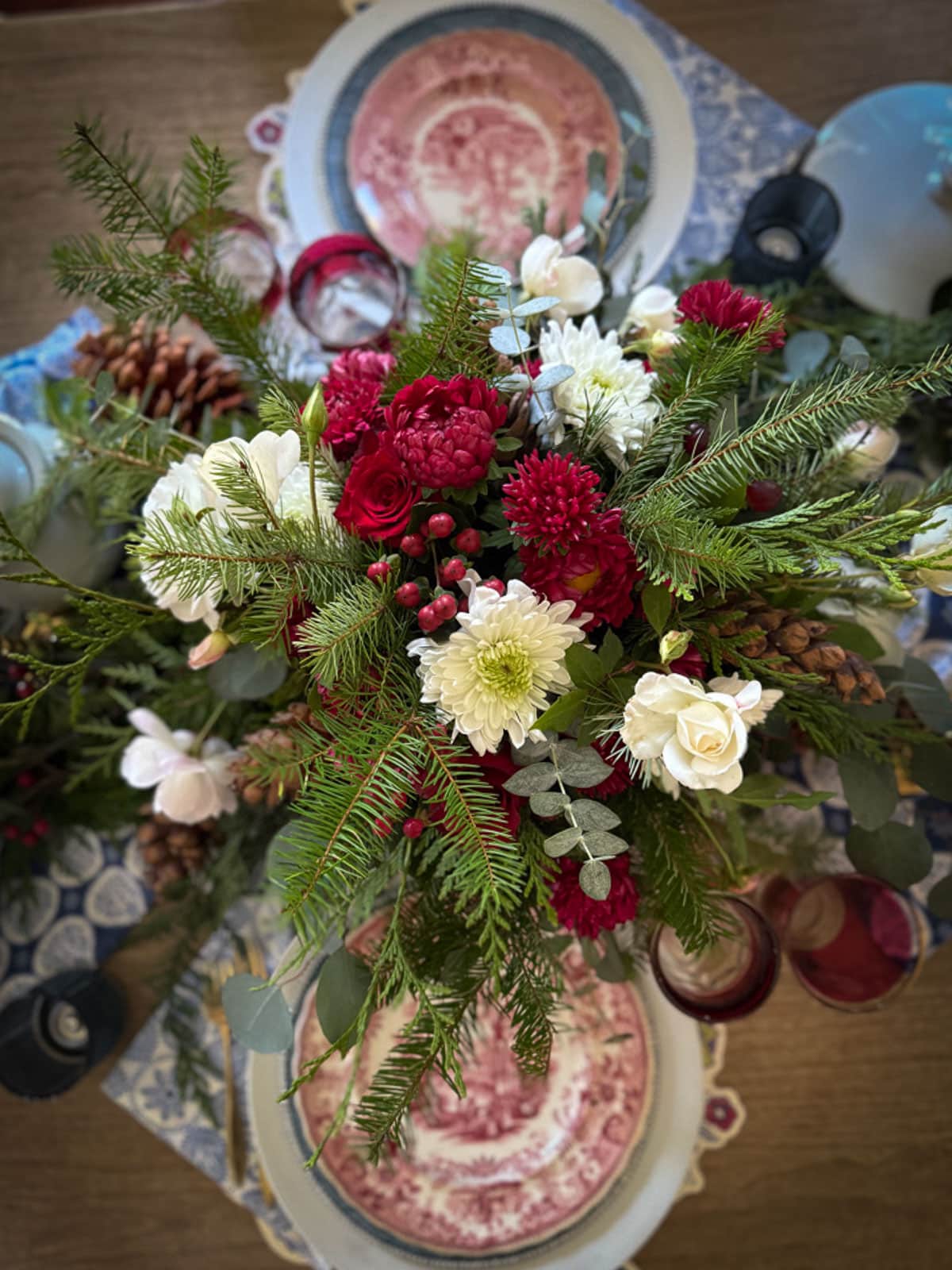 Top view of a festive table setting with a floral centerpiece. The arrangement includes red and white flowers, evergreen branches, and pinecones. Plates with red designs, glassware, and a patterned tablecloth are visible beneath the centerpiece.