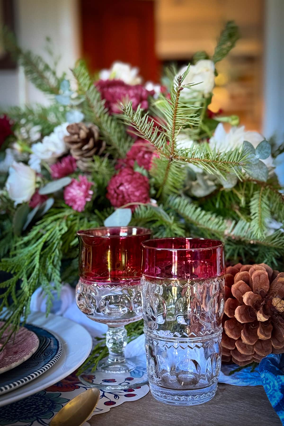 Festive table setting featuring red and clear glasses, a floral centerpiece with pine branches, pinecones, and white and red flowers. Embracing classic Christmas decor, there is also a decorative napkin and a plate, creating a cozy winter holiday atmosphere.