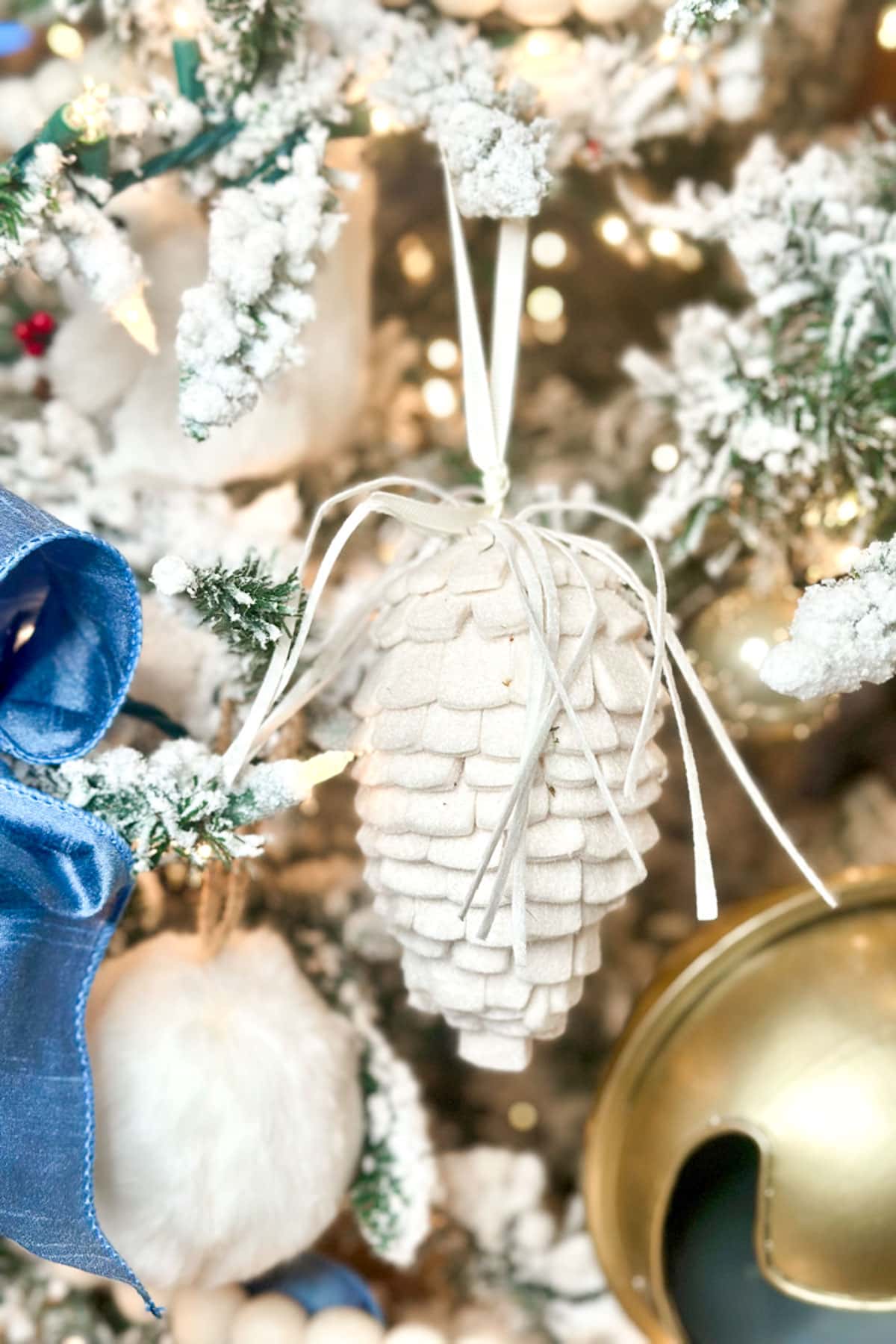 Close-up of a Christmas tree branch decorated with a white pinecone ornament tied with a ribbon. The branch is frosted with artificial snow. Nearby, a large gold ornament and a blue ribbon are partially visible.