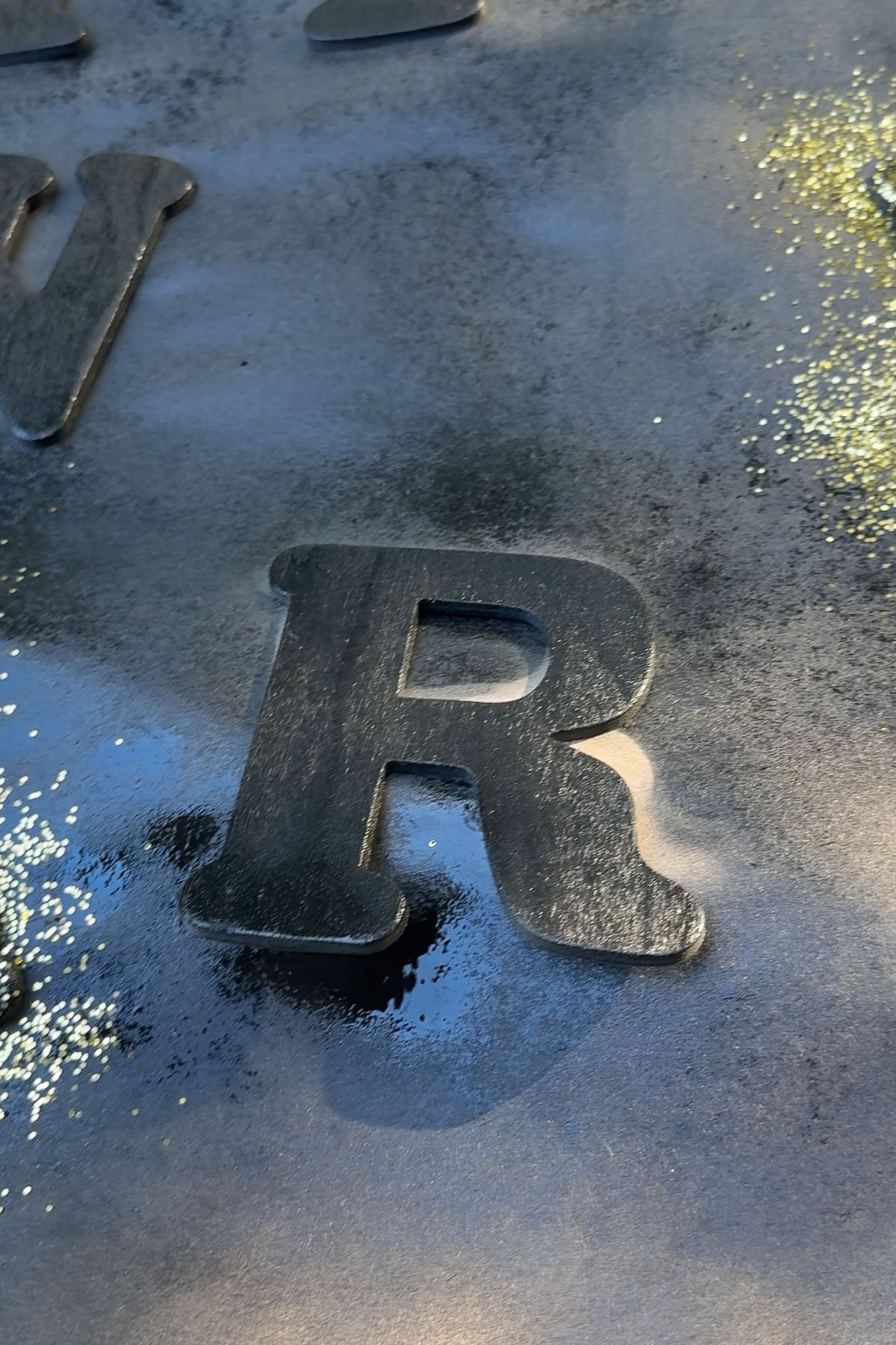 Close-up of a metallic letter "R" on a textured surface with scattered gold and black paint splatters. The sunlight creates a slight reflection on the metallic surface.