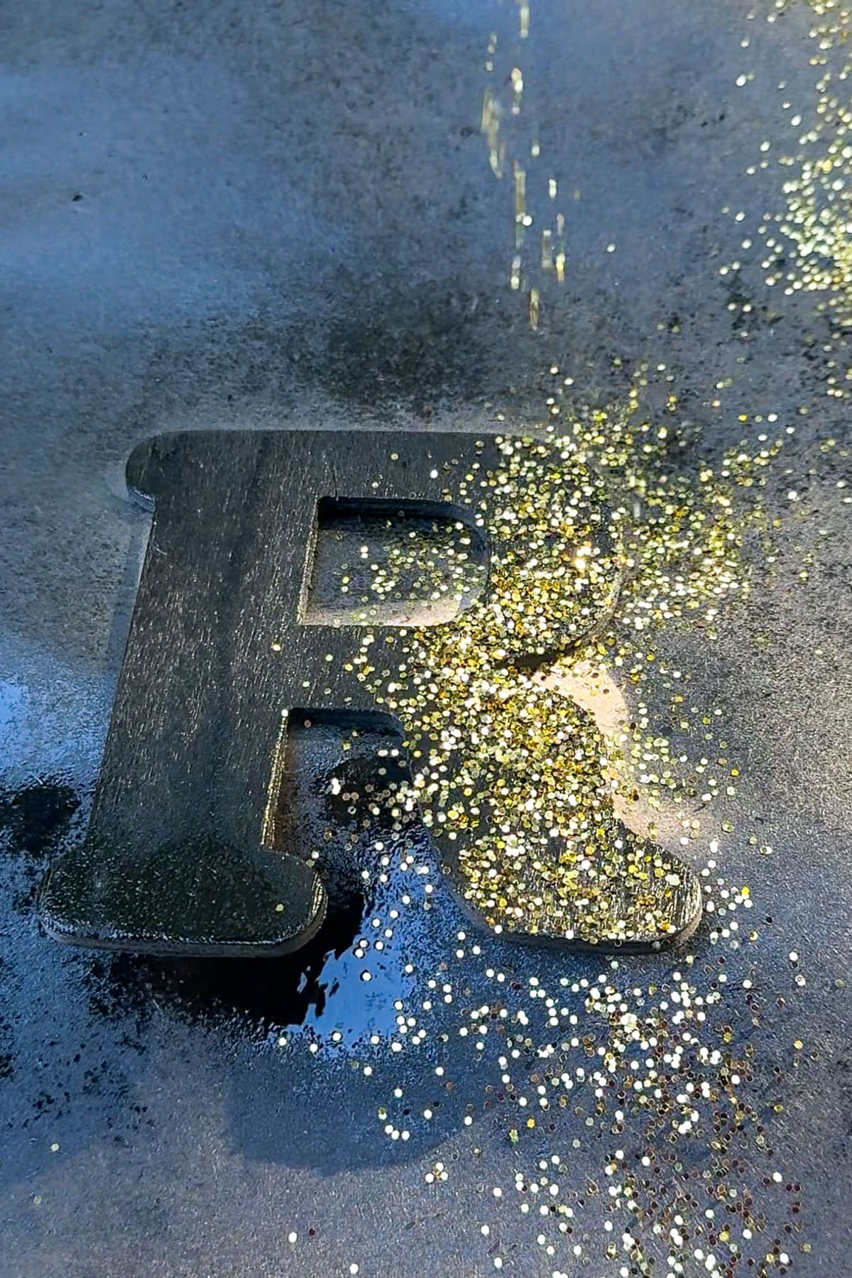 A block letter "R" on a dark surface is partially covered with scattered gold glitter. Light reflects off the glitter, creating a shimmering effect around the letter.