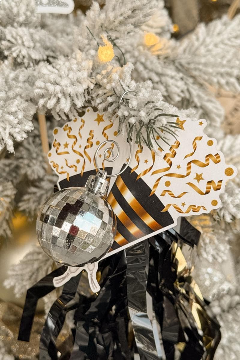 A Christmas tree branch adorned with a small silver disco ball ornament, a festive gift tag with black and gold confetti design, and black and gold metallic streamers. The tree is dusted with white snow and soft yellow lights.