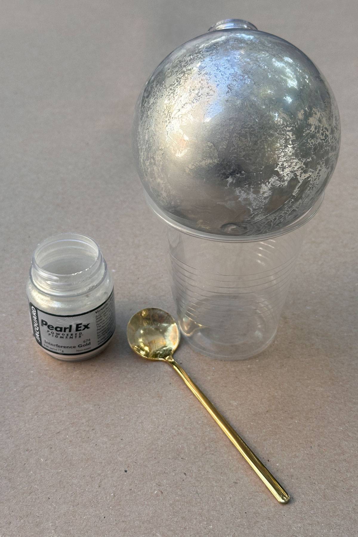 diy mercury glass ornaments-A container of Pearl Ex silver pigment, a gold-colored spoon, and a clear cup with a silver-colored orb resting on top, partially covered in the pigment. The items are placed on a beige surface.